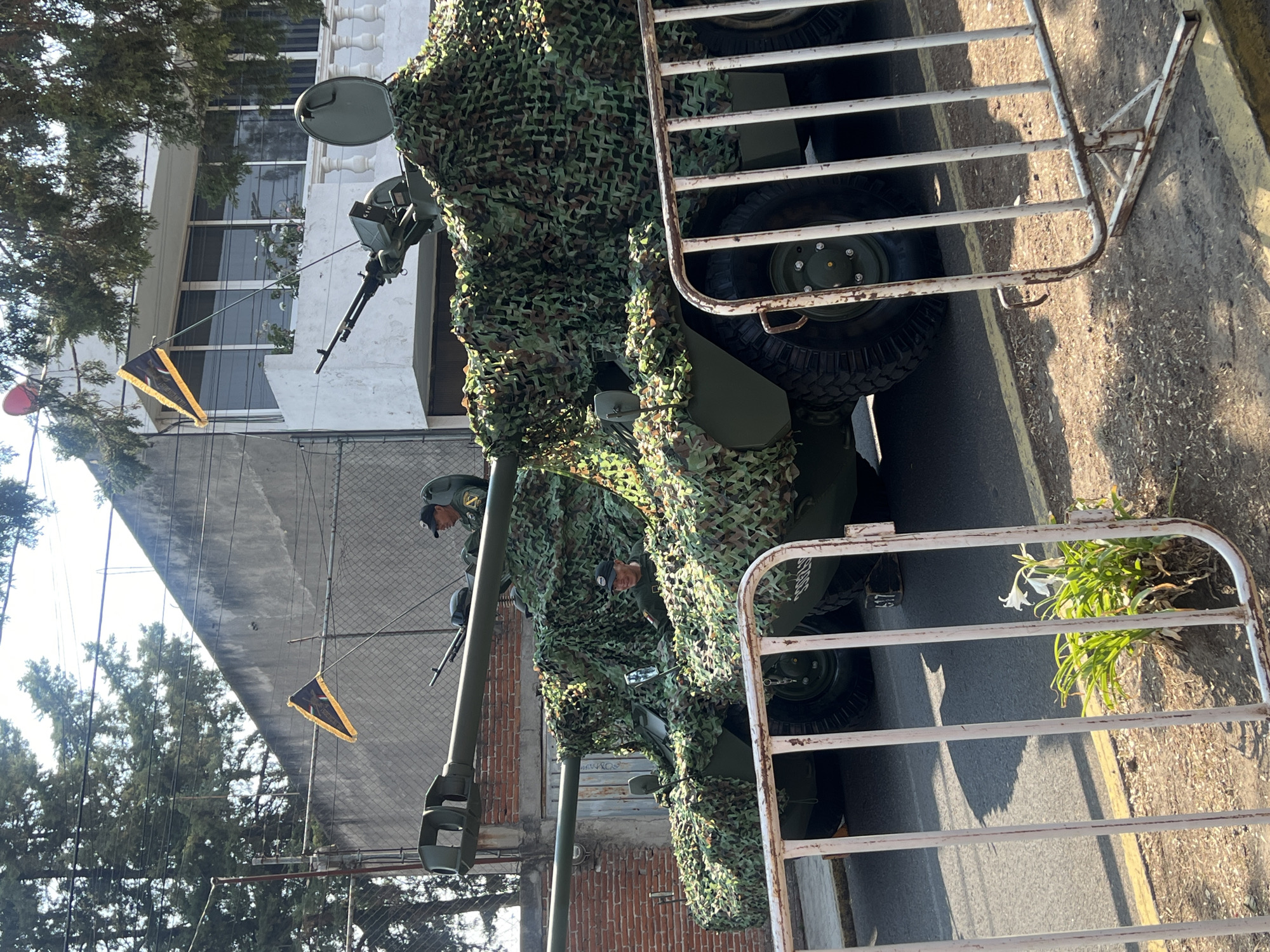 The image depicts a military vehicle covered in camouflage netting parked on a city street. The street is lined with white metal crowd control barriers on one side, with a building featuring white walls, windows, and a balcony visible in the background. A military person wearing a uniform is visible behind the camouflage netting, possibly operating the vehicle's mounted weapon. Triangular flags hang from a wire above the vehicle. The location is Unknown, Unknown.