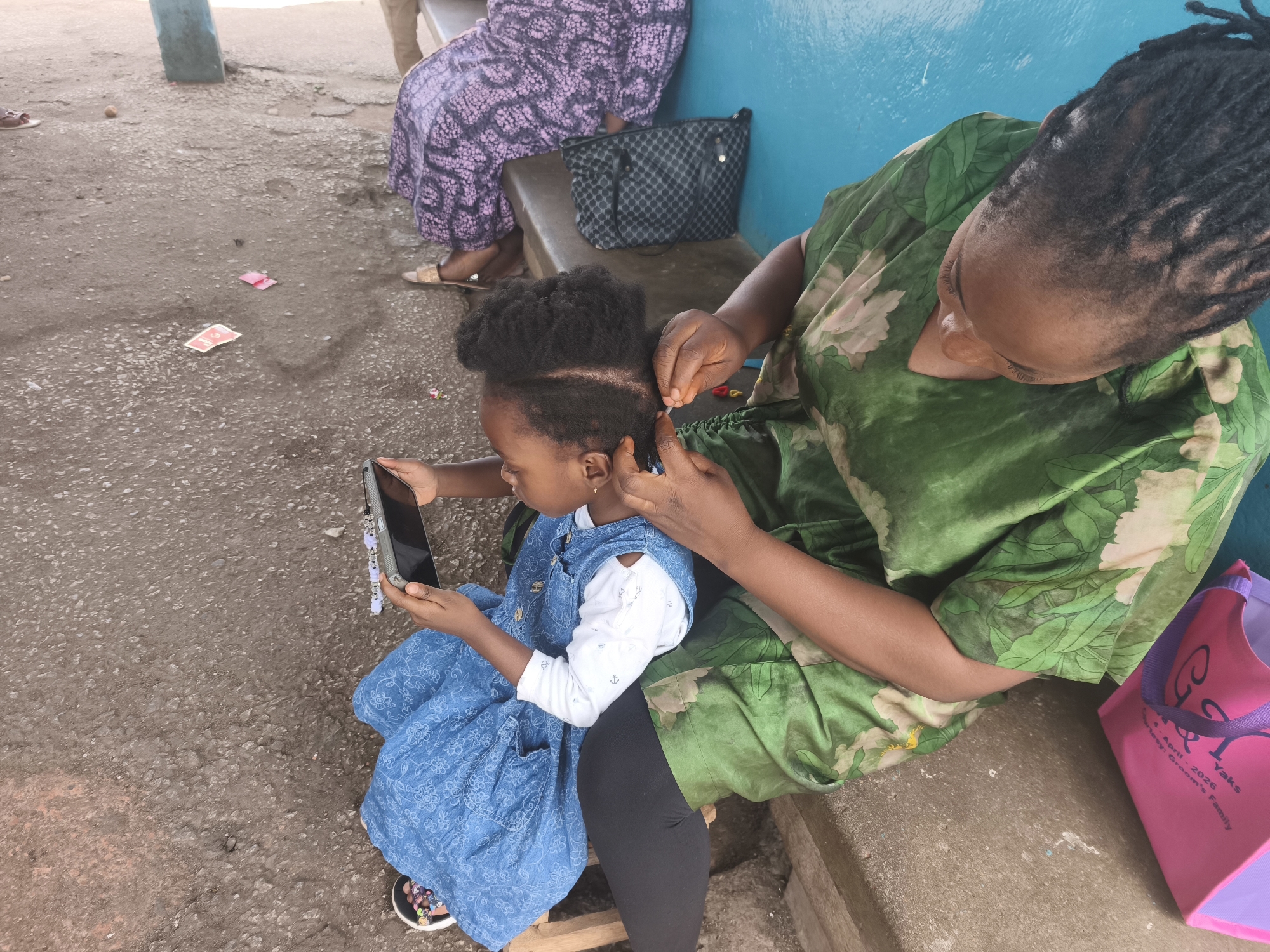 The image captures an outdoor scene in what appears to be a casual, everyday setting, identified as Jos, Nigeria. The primary focus is on two individuals: an adult woman and a young child. The woman, with her back mostly to the camera, is seen meticulously braiding the child's hair. She wears a vibrant green fabric with a lighter green floral or leafy pattern and has dreadlocks.
The child, seated in front of the woman and facing towards the left of the frame, is wearing a blue denim-like overall dress over a white long-sleeved top adorned with small anchor motifs. Her natural black hair is being worked on, with some braids already formed and others in progress. She is holding a black smartphone with both hands, seemingly engrossed in its content, suggesting a calm and routine activity.
In the background, another person is partially visible, seated, wearing a long purple patterned skirt and sandals. Behind them, a black patterned handbag rests on a bench or low wall. The setting features a blue-painted wall and a dirt or gravel ground. To the right, a pink and purple reusable bag is visible, with text partially readable as "4 April 2026," "Yaks," and "Courtesy: Groom's Family," indicating it might be a souvenir from an event. The overall ambiance suggests a relaxed daytime environment, focusing on a personal care moment between the woman and child.