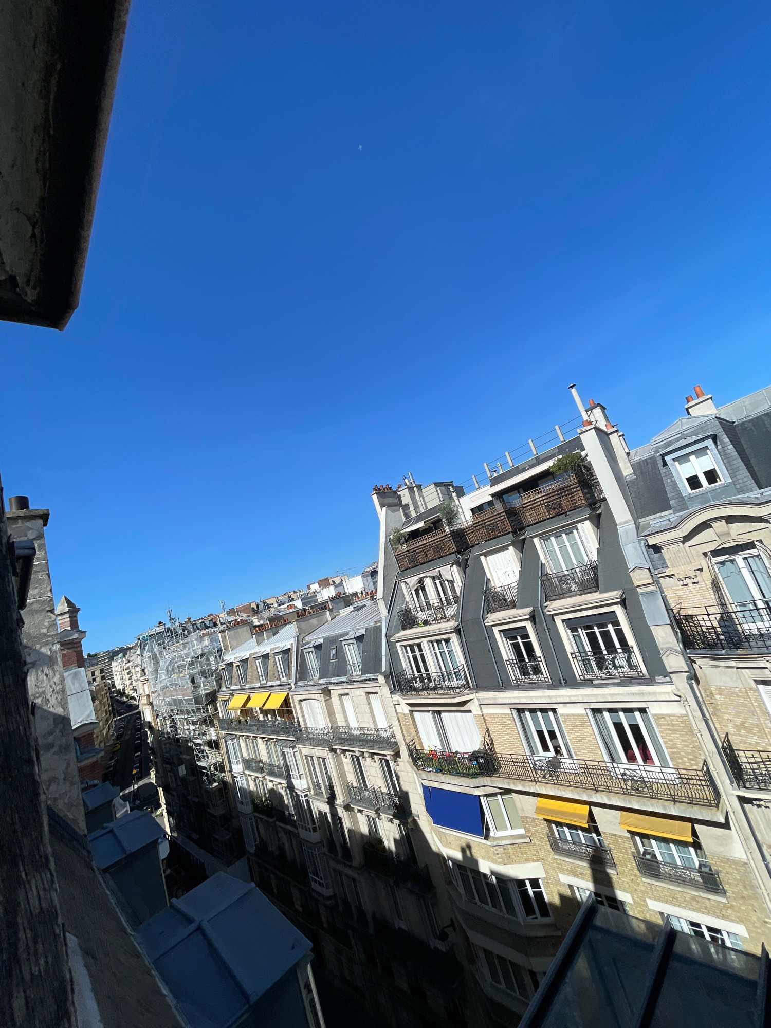 The image captures a view of Parisian buildings from a high vantage point, likely a balcony or rooftop, looking down a narrow street lined with Haussmannian architecture. The sky is a clear, vibrant blue. The buildings are characterized by their grey mansard roofs, cream-colored stone facades, and numerous windows, many with Juliet balconies featuring black wrought-iron railings. Some windows are open, revealing glimpses of interior spaces, and a few balconies have potted plants or small tables. Several buildings have yellow awnings extended over windows, and one building features a striking blue awning. Scaffolding is visible on one of the buildings, suggesting ongoing renovation or construction. The perspective is slightly tilted, emphasizing the depth and density of the urban environment. The scene is devoid of people.