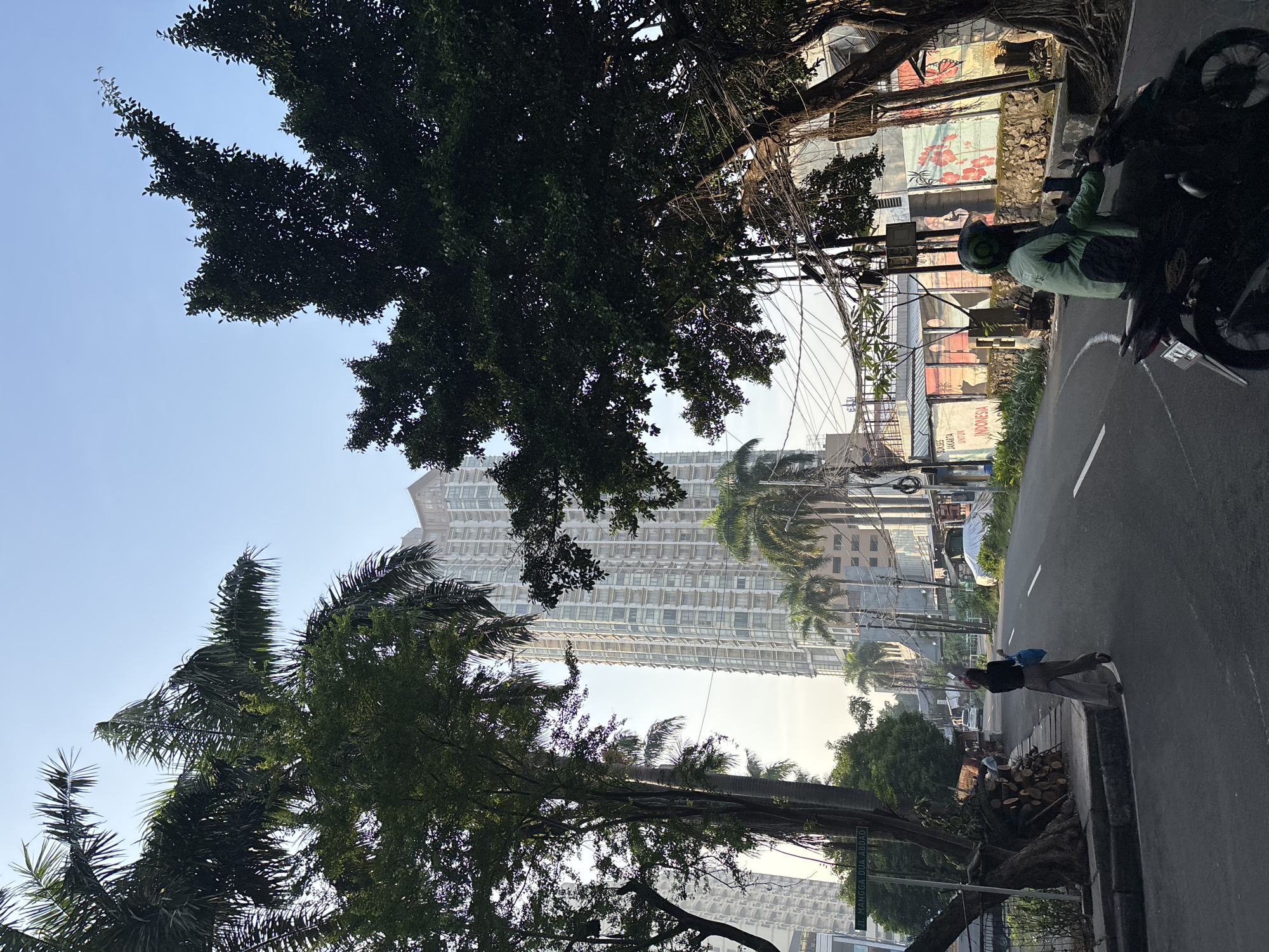 The image shows a street scene in Central Jakarta, Indonesia. The view is taken from the side of a road with a sidewalk. A tall building with many windows is visible in the background, partially obscured by trees. Palm trees are also present. A person stands on the sidewalk, holding a book and a blue bag. A motorcycle is parked on the side of the road near a building with a mural. Overhead, power lines crisscross. There is a green street sign. A pile of chopped wood sits on the sidewalk.