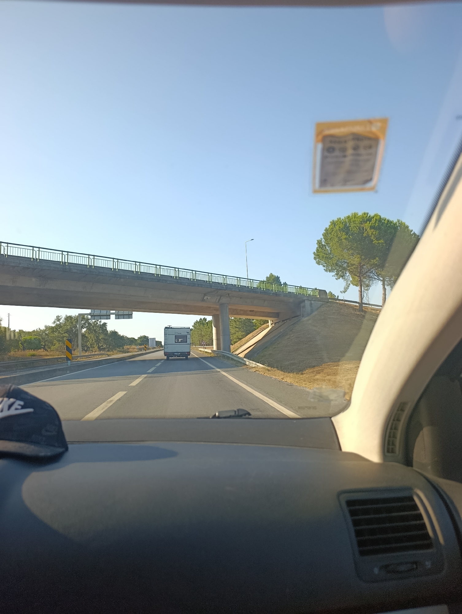 The image captures a view from inside a vehicle on a highway, likely in or near Alcácer do Sal, Portugal. The road ahead has multiple lanes and is bordered by dry grass and trees. A large concrete overpass with a green railing spans the highway in the mid-ground. A white recreational vehicle (RV) is visible driving away from the camera on the highway, passing under the overpass. A signpost with directional information is mounted on the overpass. On the right side of the frame, a tall, mature pine tree stands on a grassy embankment. The sky is clear and blue, suggesting a sunny day. A black baseball cap with a white logo is partially visible on the dashboard in the lower left corner of the image. A yellow sticker is affixed to the windshield. The interior of the vehicle, including the dashboard and air vents, is also visible.