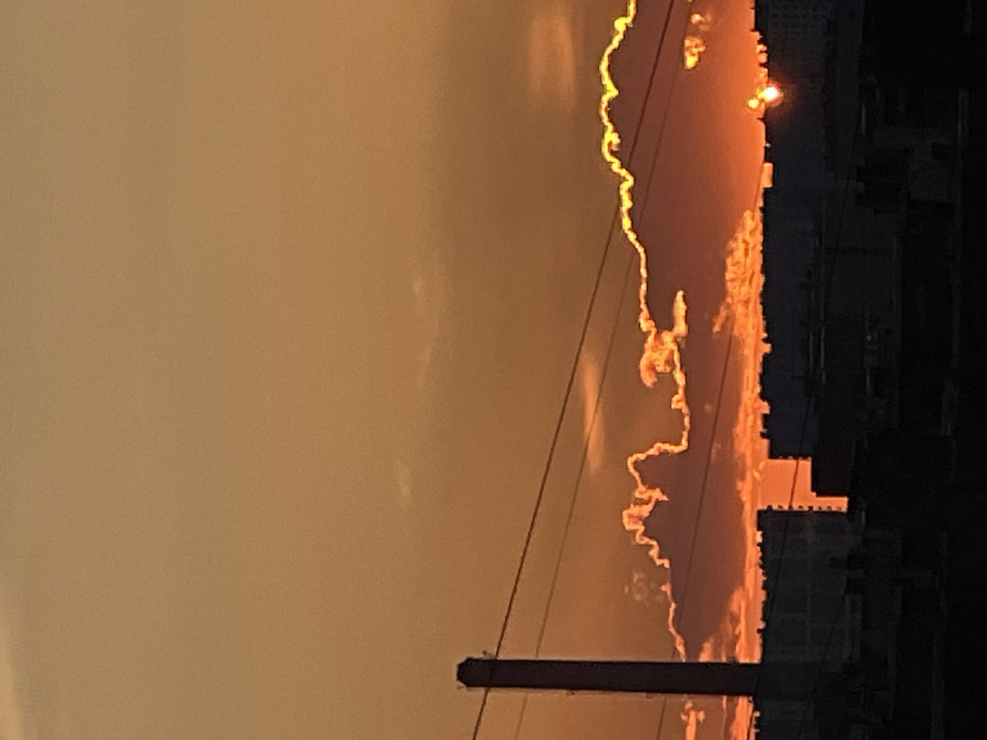 The image captures a sunset over the city of Ladozhskoe Ozero, Russia. The sky is a warm gradient of orange and yellow, transitioning to a lighter hue at the top. Silhouetted against the vibrant sky are the outlines of buildings and a tall industrial chimney on the left. Power lines traverse the mid-ground horizontally. Fluffy clouds are illuminated from below by the setting sun, creating a dramatic effect with their edges glowing in golden and orange light. The sun itself is a small, bright orb peeking through a gap in the clouds above the buildings. The lower portion of the image is dominated by the dark silhouettes of urban structures.
