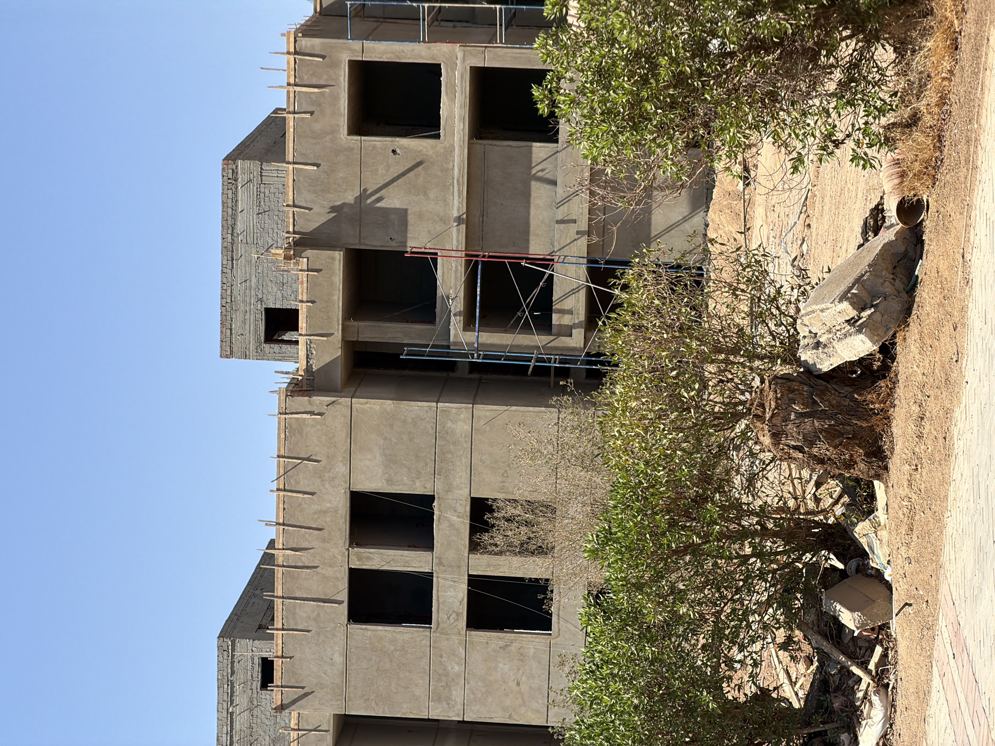 The image shows a partially constructed building in Sharm El Sheikh, Egypt. The building is made of concrete and appears to be several stories high. Some of the windows are visible as dark openings, and scaffolding is present on the exterior. The construction appears to be in an early to mid-stage, with wooden beams and rebar exposed in places.
Surrounding the building is an arid, sandy landscape with sparse vegetation. A tree with green leaves is prominently featured in the foreground, partially obscuring the building. In the lower right of the image, there is debris, including concrete fragments and what appears to be trash. A paved pathway runs along the edge of the scene.
The lighting suggests it is daytime, likely mid-morning or afternoon, with clear, bright sunlight casting distinct shadows. The sky is a pale blue, indicating fair weather. The scene conveys a sense of ongoing construction and development in a warm, dry climate, common to the region of Sharm El Sheikh. There is no visible text in the image.