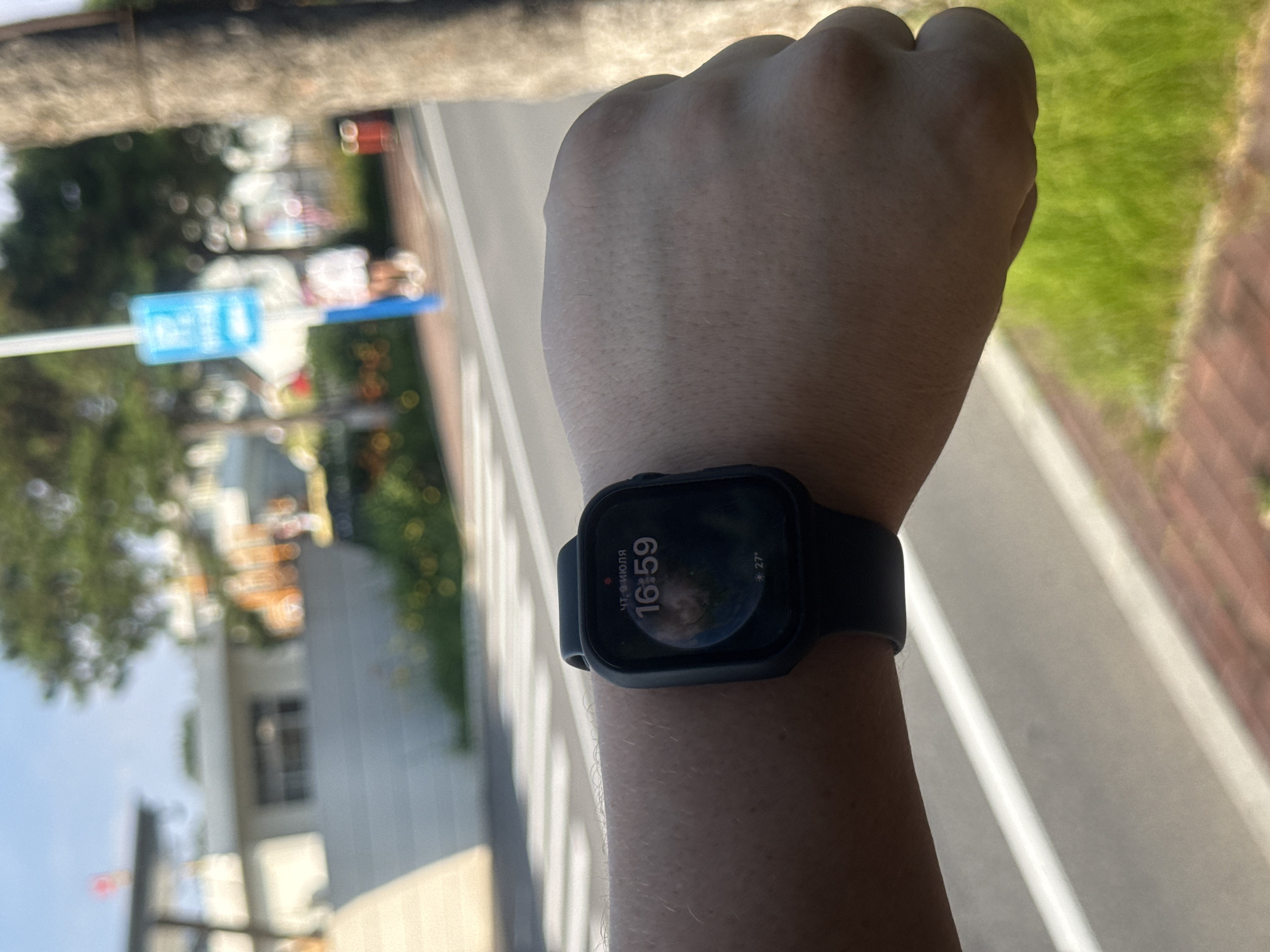 A first-person perspective shot shows a person's left arm and clenched fist, wearing a black Apple Watch. The watch face displays the time as 16:59 and the date as "ЧТ, 9 ИЮЛЯ" (Thursday, July 9th). It also shows the temperature as 27 degrees Celsius. The background is blurred but appears to be a street scene. A pedestrian crossing is visible in the foreground, and a tree trunk is on the right side of the frame. In the distance, buildings and trees are visible. The environment suggests an outdoor, urban setting in Qinhuangdao, China, based on the date format and likely location of such a scene.