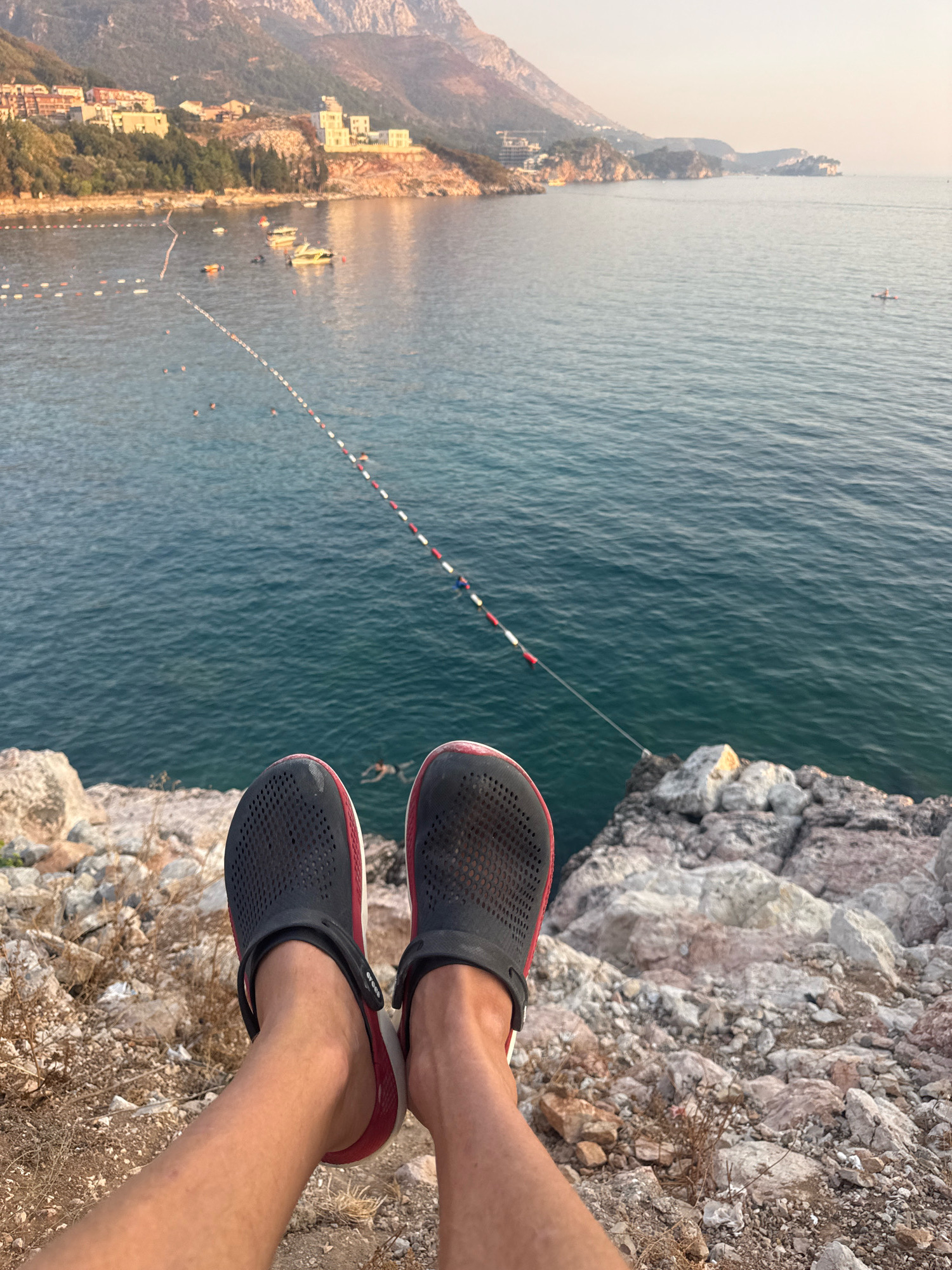 The image captures a scenic view of the Adriatic Sea from a rocky outcrop in Kamenovo, Montenegro. In the foreground, a pair of black Crocs with red accents are worn by a person whose legs and feet are visible, resting on the rocky terrain. The person is positioned to overlook the sea, which is a deep blue color and exhibits slight ripples. A bouy line with red and white markers stretches across the water, delineating a swimming area. Several boats, including a yellow yacht, are anchored or moving in the distance. On the shore, buildings with red-tiled roofs are nestled among lush green trees, with a mountainous landscape forming the backdrop. The sky is a soft, hazy light, suggesting either early morning or late afternoon. A single person can be seen on a paddleboard in the middle of the sea.
