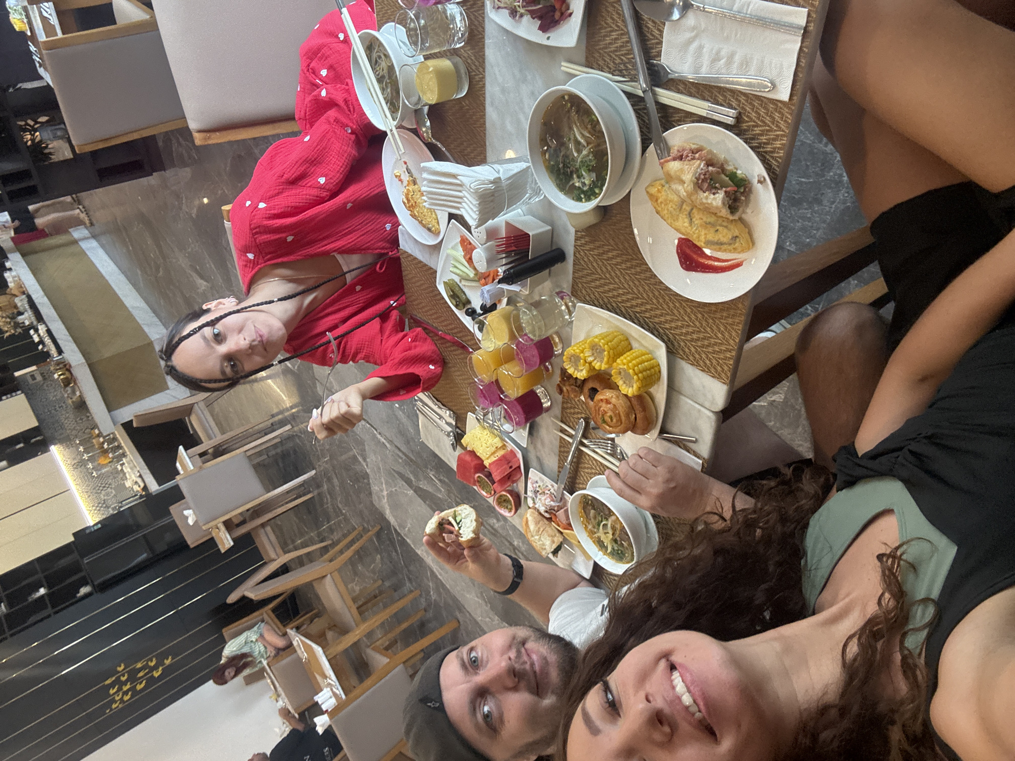 Four individuals are seated around a table laden with food, presumably at a restaurant, captured in a selfie-style shot. The individuals are facing the camera, partially visible from their torsos up. The focus is on the foreground, where two people are taking the selfie.
The table displays a variety of dishes, including bowls of soup, plates with omelets or wraps, corn on the cob, and what appears to be cut-up fruit. There are also several glasses of liquids, possibly juice, and a stack of napkins. Silverware and chopsticks are visible among the dishes.
The environment suggests a dining area, with visible furniture like chairs and a countertop, as well as elements of interior design, such as a patterned floor and wall decorations. The location is identified as Đà Nẵng, Vietnam.