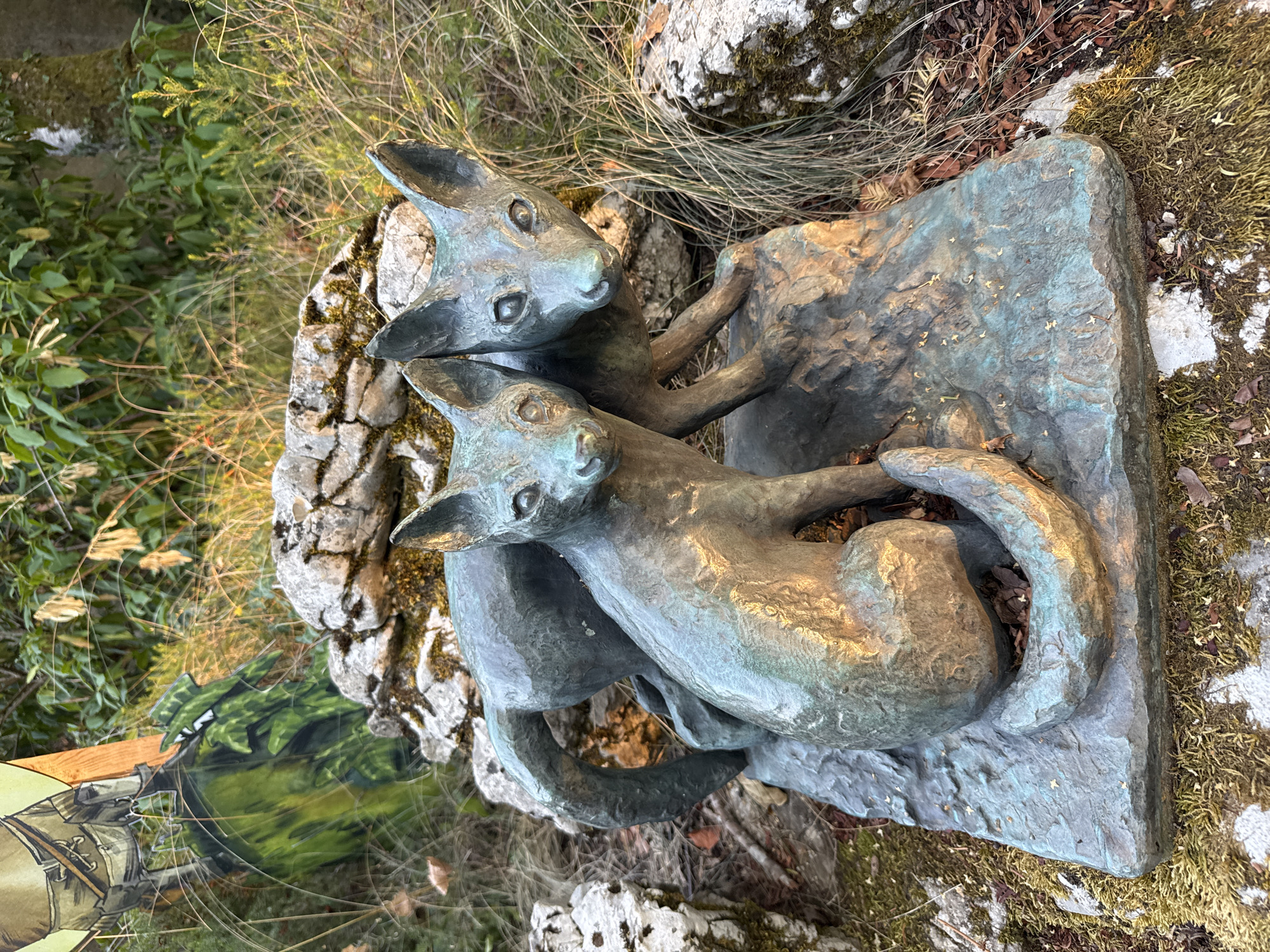A bronze sculpture of two foxes is positioned outdoors, nestled amongst rocks and dry grass. The sculpture is situated on a mossy, uneven surface that appears to be part of the natural environment. Behind the foxes, a large, rough-hewn rock forms part of the background. To the left of the sculpture, a portion of a flat, colorful image is visible, depicting a figure in green camouflage clothing and what appears to be part of a soldier's uniform. The scene is illuminated by natural light, casting shadows and highlighting the metallic patina of the sculpture. The overall setting suggests an outdoor display or installation, possibly in a park or garden. While the image does not provide specific clues to its exact location within Porrentruy, Switzerland, the naturalistic setting of rocks, grass, and foliage is consistent with many outdoor areas in the region.