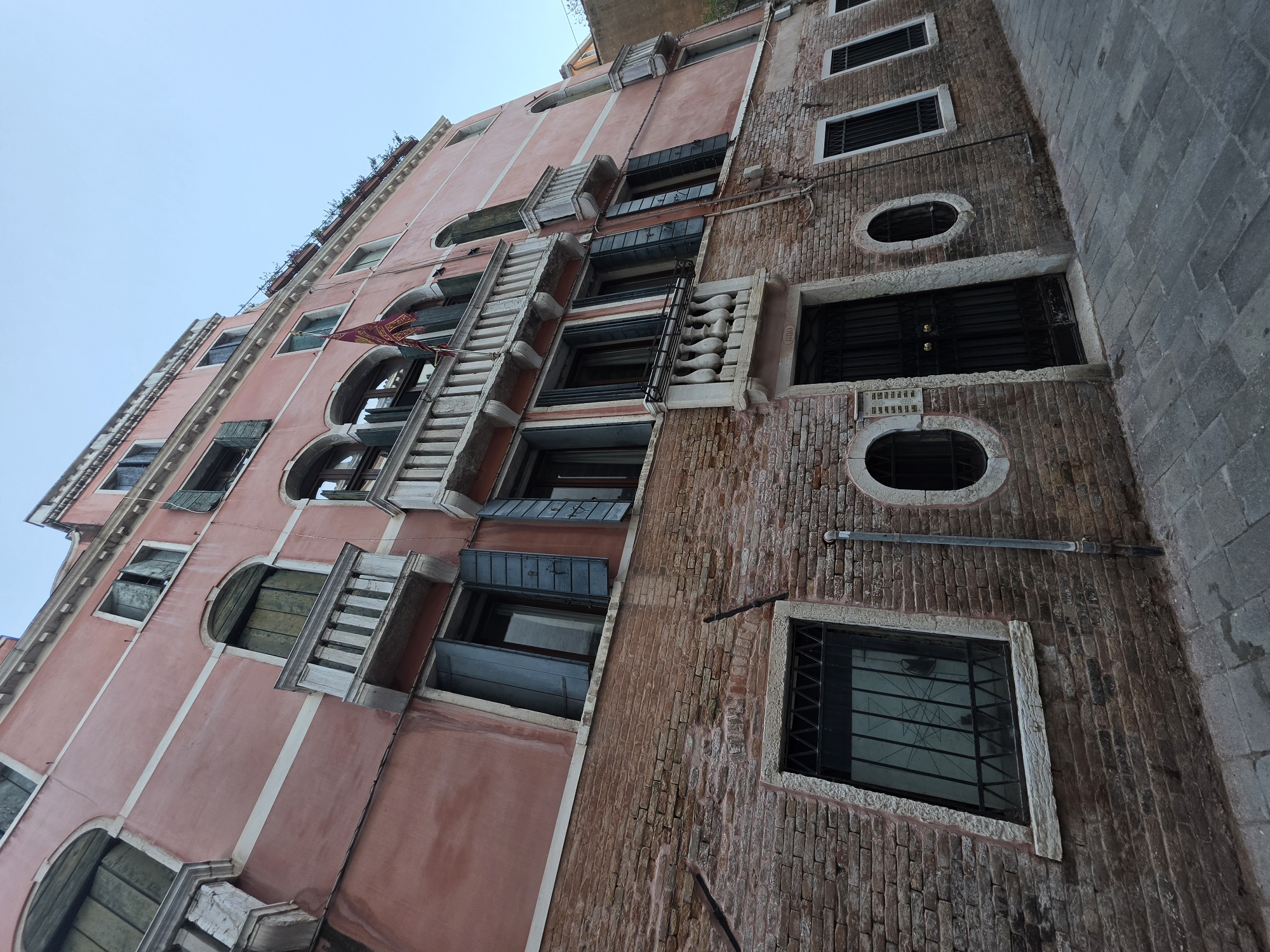 This is a low-angle shot of a brick building in Venice, Italy. The building is pink with white trim and features several arched windows with green shutters. Some windows have small balconies with ornate railings. A red and gold flag hangs from one of the balconies. The building's lower section is made of weathered red brick, and it has a dark metal door and several barred windows. The ground in front of the building is paved with cobblestones. The sky is clear and blue.