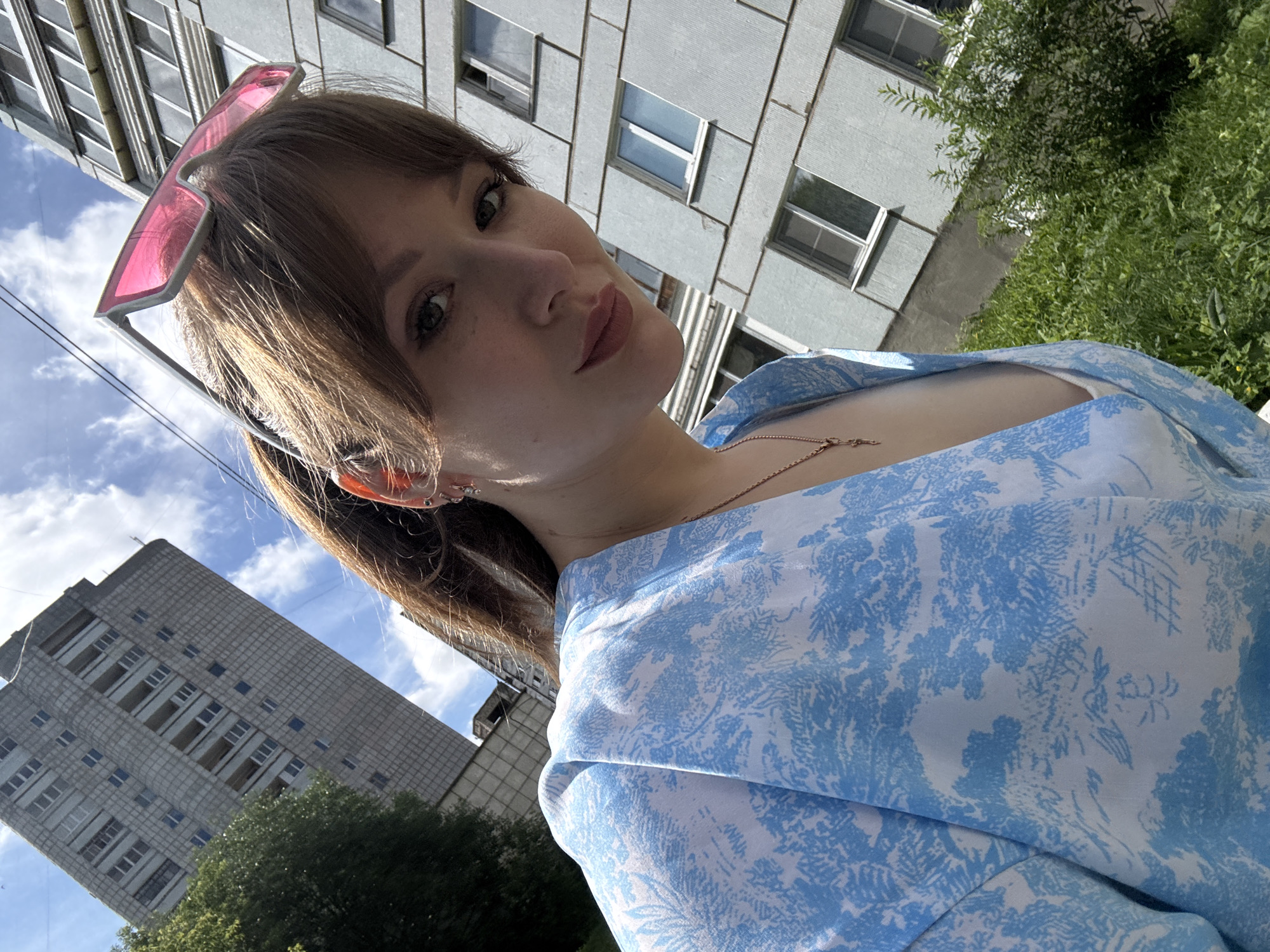 A young woman with brown hair and bangs, wearing pink sunglasses perched on her head and a blue and white patterned dress, takes a selfie outdoors. She is looking at the camera with a slight smile. The background features a grey, multi-story building with numerous windows, a tall residential building in the distance, and green trees and bushes. The scene appears to be set in a city, possibly in Chaikovsky, Russia, given the architectural style of the buildings. The sky is blue with white clouds.