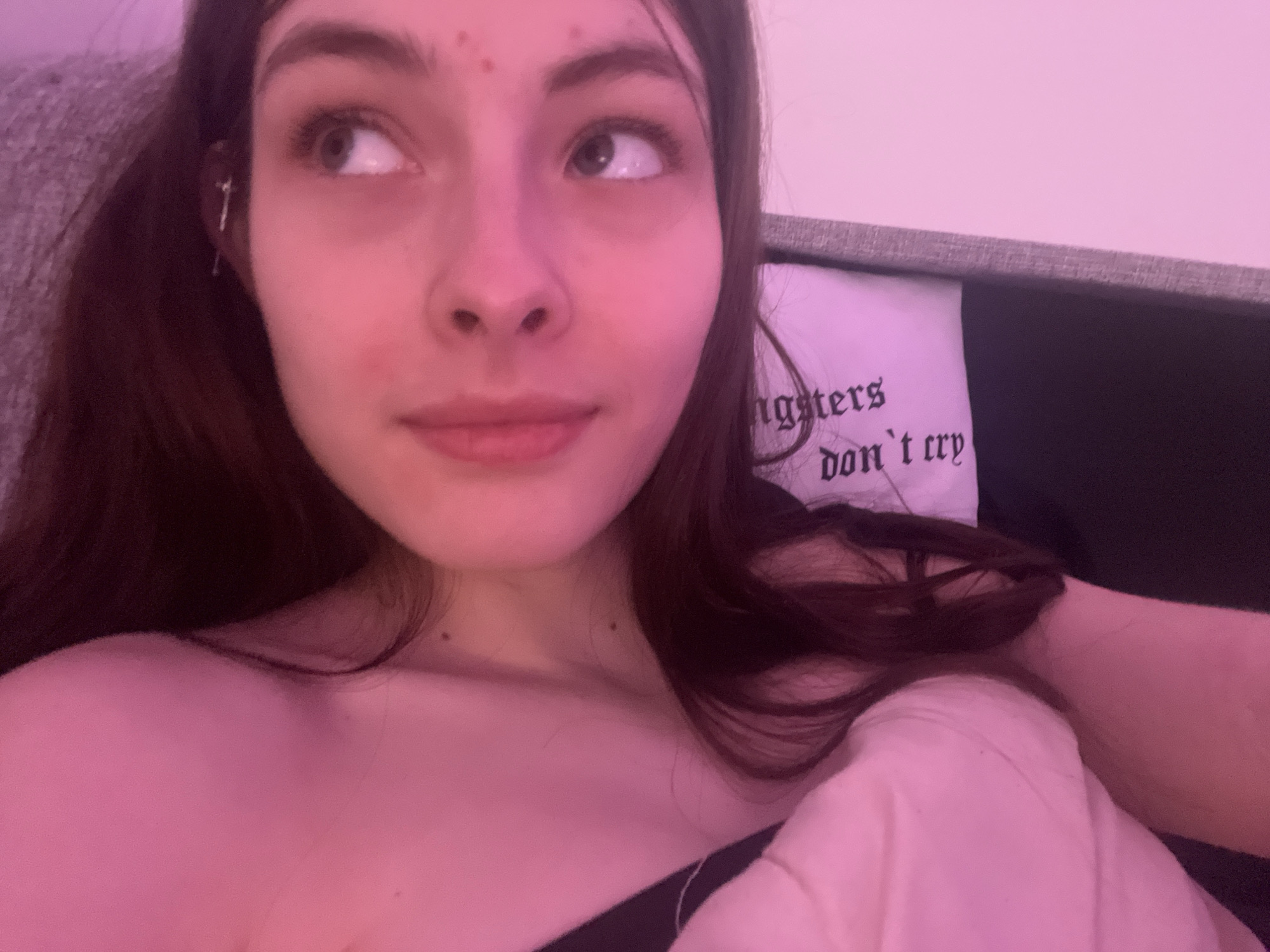 A close-up of a fair-skinned young woman with long brown hair lying down, possibly on a sofa or bed. She has dark eyebrows and light-colored eyes, and a few blemishes on her forehead and cheek. She is looking to the left of the frame. She wears an earring. A white pillow or cushion with the inscription "gangsters don't cry" is partially visible behind her head. A blanket is partially obscuring the subject's chest. The lighting appears to be pinkish or purplish, giving the image an overall warm tone. The location in Saint Petersburg, Russia, cannot be determined based solely on the content of the image.