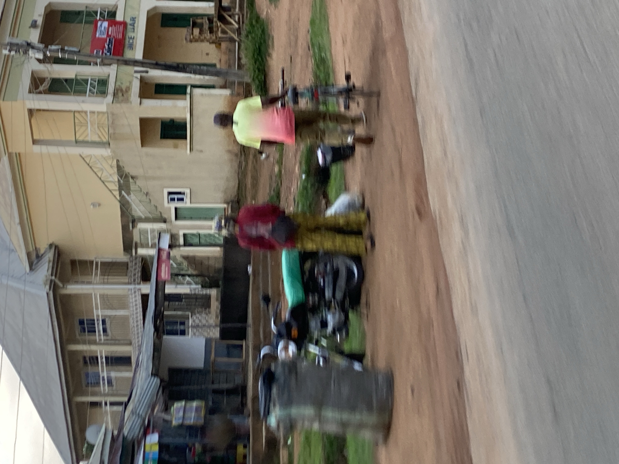 In a street scene in Jos, Nigeria, two individuals are observed near a roadside. To the left, a person wearing a red jacket and yellow checkered pants stands near a black motorcycle with a bright green seat. A large, cylindrical gray object is positioned to the far left of the motorcycle. To the right, another person, seen from the back, is wearing a shirt that transitions from yellow at the top to pink at the bottom, and brown pants. This individual is standing next to a bicycle. In the background, a two-story building with light yellow and green accents is visible, featuring an external staircase and balconies. A sign on the building reads "ICE BAR". Shops with corrugated metal roofs are present to the left of the building. The ground is a mix of dirt and asphalt, with some sparse green vegetation.
