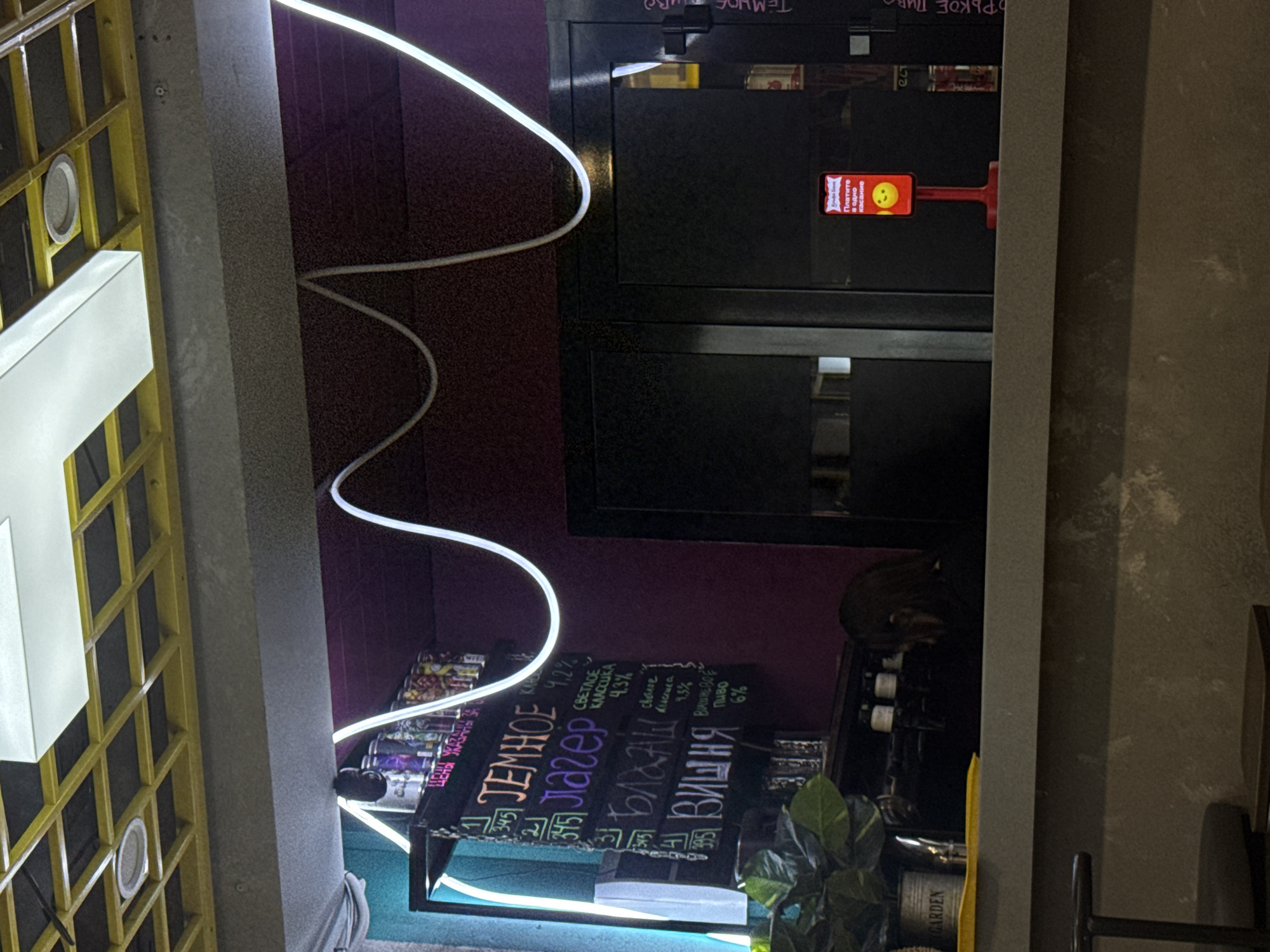 The image depicts an indoor setting. There's a vertical panel covered in a yellow lattice design to the left. Next to it, a grey concrete pillar extends upwards. In the middle, there's a dark purple wall with a black double door. The lower door appears to be open, revealing some shelves and bottles. In front of the wall and door, a chalkboard displays the prices of beverages, including "TEMHOE," "JAIEEP," "BARAH," and "BWWHYA." A white neon light fixture zigzags in front of the wall and door. A plant is placed near the chalkboard. A second concrete pillar appears to the right of the doors and wall. The location is believed to be Mozhvai, Russia.