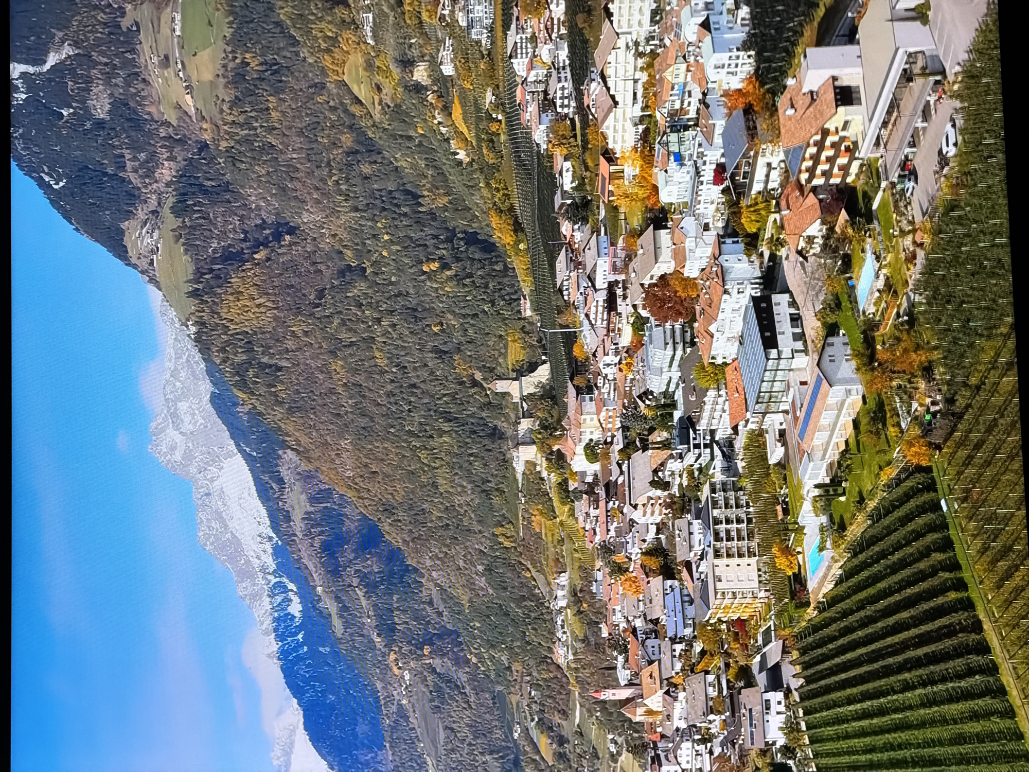 This image depicts a panoramic view of a mountainous region with a town nestled in the valley. The mountains in the background are snow-capped at the highest peaks, with lower slopes covered in dense evergreen forests and patches of deciduous trees displaying autumn colors of yellow and orange. A town with a mix of residential buildings, some with red-tiled roofs, is spread across the lower slopes and the valley floor. A prominent church with a red spire is visible in the town. In the foreground, there are rows of green trees, likely vineyards or orchards, leading down to a more developed area with modern buildings, a swimming pool, and a parking lot with several cars. The sky is a clear blue with a few wispy clouds. The scene suggests an autumn day in a European alpine setting.
This image is not of Chirchik, Uzbekistan. The architectural style of the buildings, the type of vegetation, and the mountainous landscape are characteristic of regions in the Alps or similar European mountain ranges, not Central Asia.