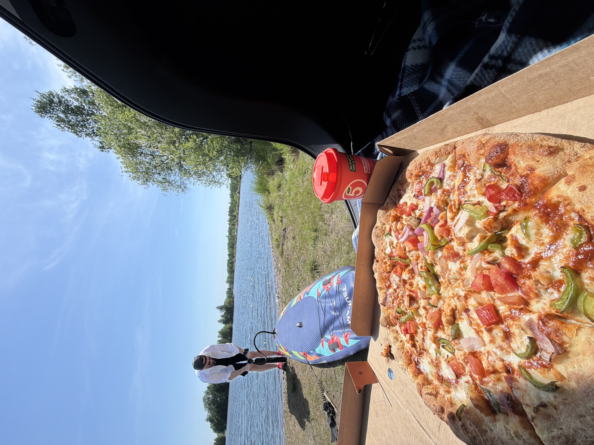 The scene is a picnic setup by a body of water. A person is inflating a blue and red paddleboard with a hand pump. To the right, a partially open pizza box reveals a pizza with red onions, green peppers, and diced tomatoes. Next to the pizza, there's a red thermos with a lid. The setting appears to be a grassy area near a lake or river, with trees visible in the background. The photo seems to be taken from inside a vehicle, with a portion of the car's interior visible at the top of the frame. There is no information in the image to indicate the location is Birobidzhan, Russia.