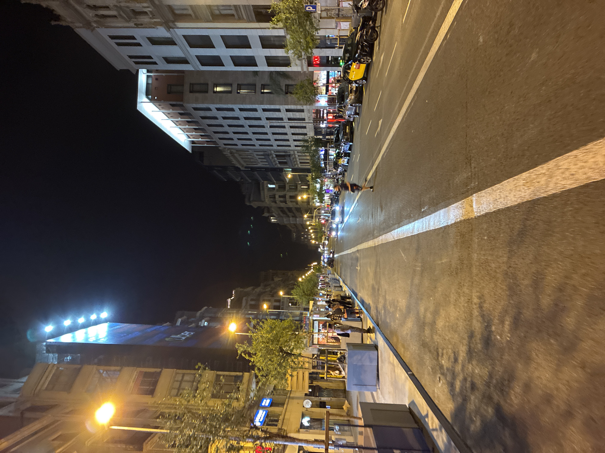 The image captures a vibrant nocturnal street scene in an urban environment, identified as Barcelona, Spain. The wide multi-lane road, possibly a boulevard, is illuminated by numerous streetlights and the glow emanating from surrounding buildings. The sky is dark, indicating nighttime.
On both sides of the road, multi-story buildings rise, their windows glowing with interior light. One building has a sign partially visible, reading "HOTEL ARK". The architecture suggests a European city, with a mix of older and more modern structures. On the right side of the street (when viewed upright), vehicles are parked, including several motorcycles and a yellow taxi, a common sight in Barcelona. Other cars are visible further down the road, with their headlights and taillights creating light streaks in the distance.
The road surface appears somewhat reflective, possibly due to recent rain or just the way the strong artificial light bounces off the asphalt. Clear white lane markings delineate the traffic flow, including a solid white line running down the center.
Pedestrians are scattered along the sidewalks, engaged in typical city night activities. One person is crossing the street. The sidewalks are lined with trees, some bus stops or advertising kiosks, and streetlights that cast warm glows. On one such kiosk, the brand name "GUERLAIN" is discernible. A blue "P" sign, commonly indicating parking, is also visible. The overall atmosphere is that of a bustling yet calm city night.