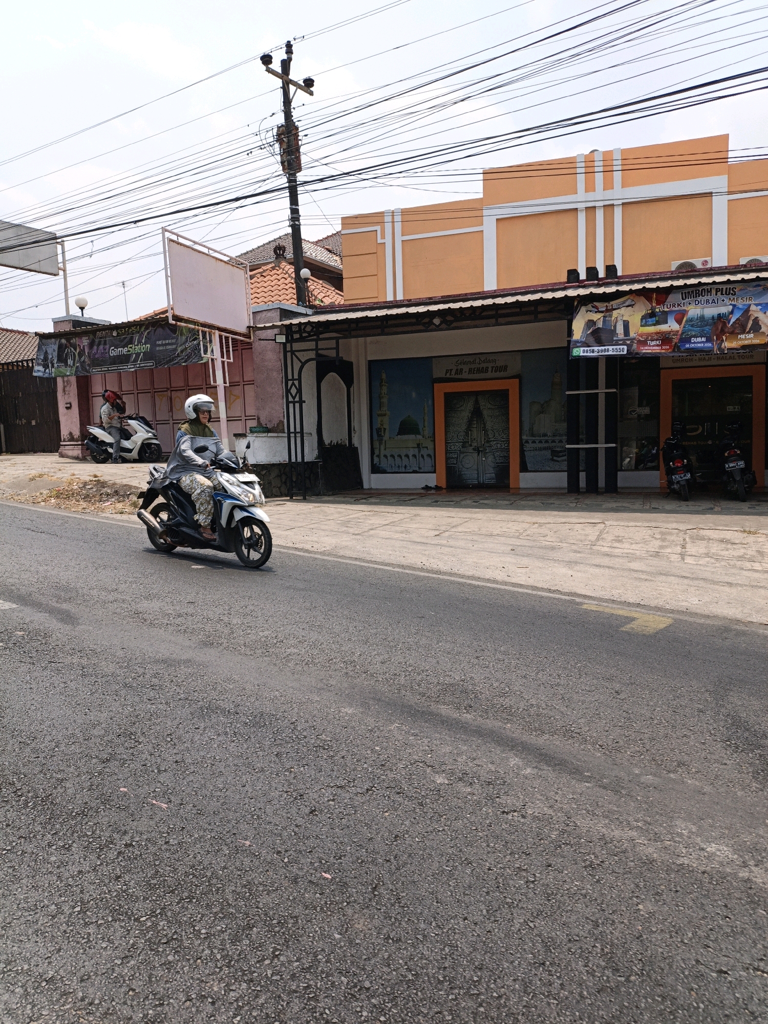 A woman wearing a white helmet and a grey shirt rides a white and blue scooter on an asphalt road in Kuwasen, Indonesia. She is wearing patterned pants. Behind her, another person is seated on a white scooter. In the background, there are buildings with orange facades and awnings. One building has signs advertising "UMROH PLUS TURKI + DUBAI + MESIR" and "PT. AR - REHAB TOUR." Another sign reads "GameStation." Utility poles and wires are visible overhead against a bright sky.