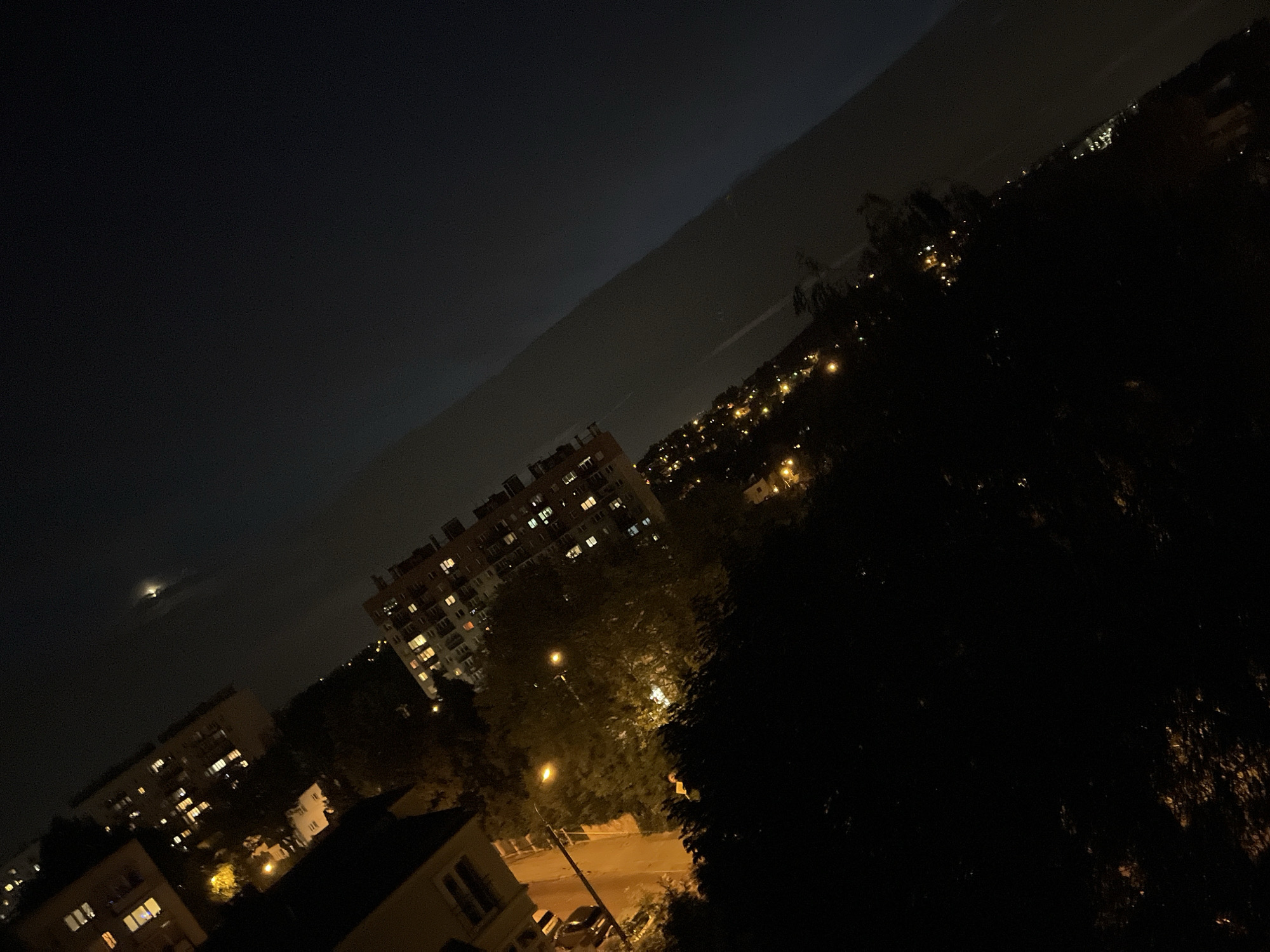 The image is a tilted night view of a city scene. In the foreground, dark foliage is visible, with some scattered yellow lights, possibly from trees or decorations. Behind the foliage, a building with lit windows is partially obscured by trees. Further back and to the left, another building with illuminated windows is visible, appearing to be residential. A street lamp illuminates the area between these buildings. In the upper portion of the image, the sky is dark with a faint glow, and a distant, bright object, possibly the moon or a satellite, is present. The horizon shows a hazy outline of distant city lights, suggesting a body of water or a larger urban area. The scene depicts a dimly lit urban environment at night, with residential buildings and natural elements.