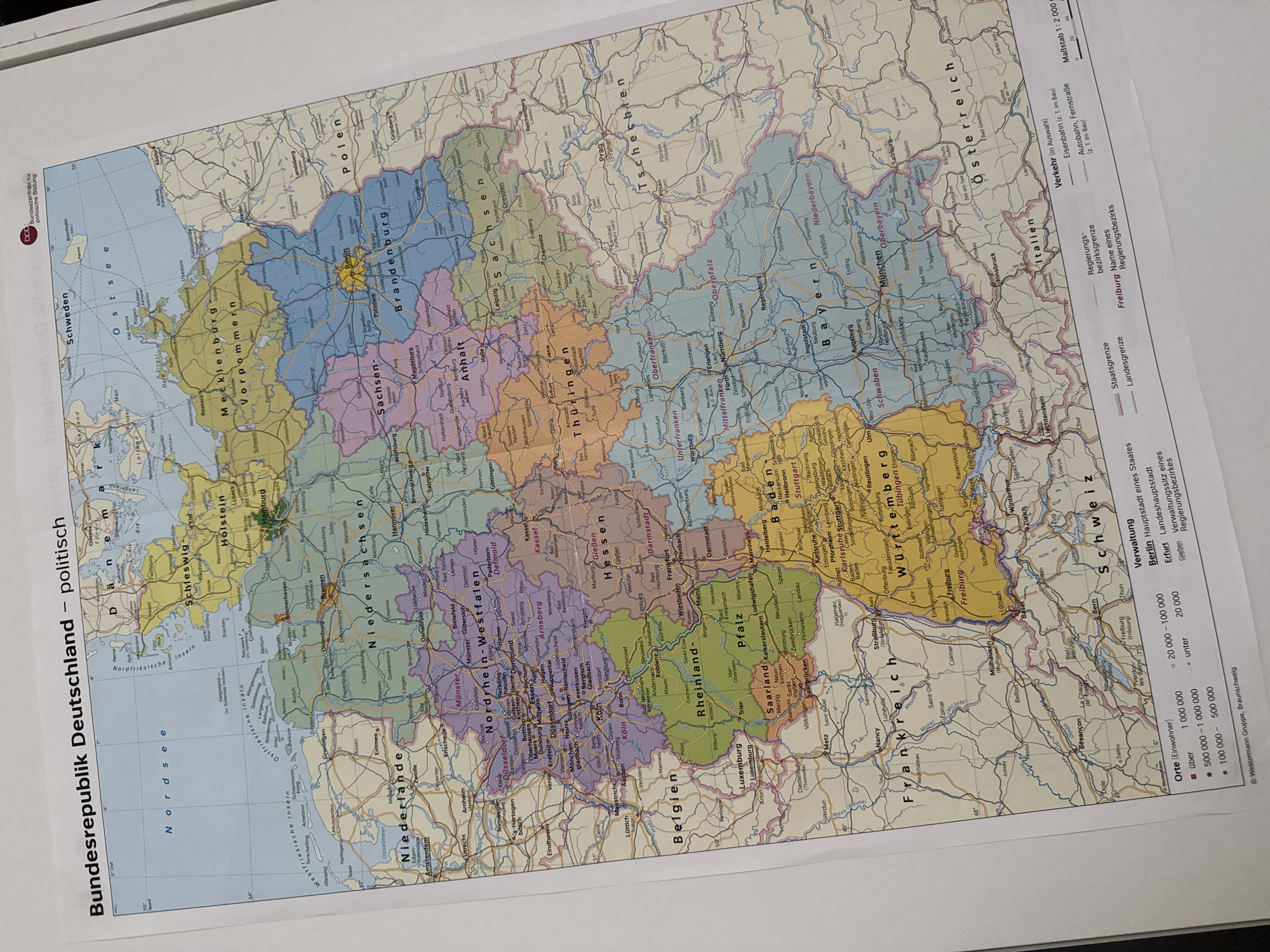 The image is a photograph of a political map of Germany. The map is titled "Bundesrepublik Deutschland – politisch" which translates to "Federal Republic of Germany – Political". It displays federal states, major cities, towns, rivers, and international borders. The map is in color, with different states represented by distinct colors. A legend in the lower right corner provides information on population density of cities, state borders, and regional districts, as well as transportation infrastructure like railways and highways. There are also scale bars indicating distances.
The city of Bad Sachsa, Germany is located on this map in the federal state of Lower Saxony (Niedersachsen), in the south-eastern part of the state, near the border with Thuringia (Thüringen). It appears as a small town represented by a dot, with its name clearly labeled.