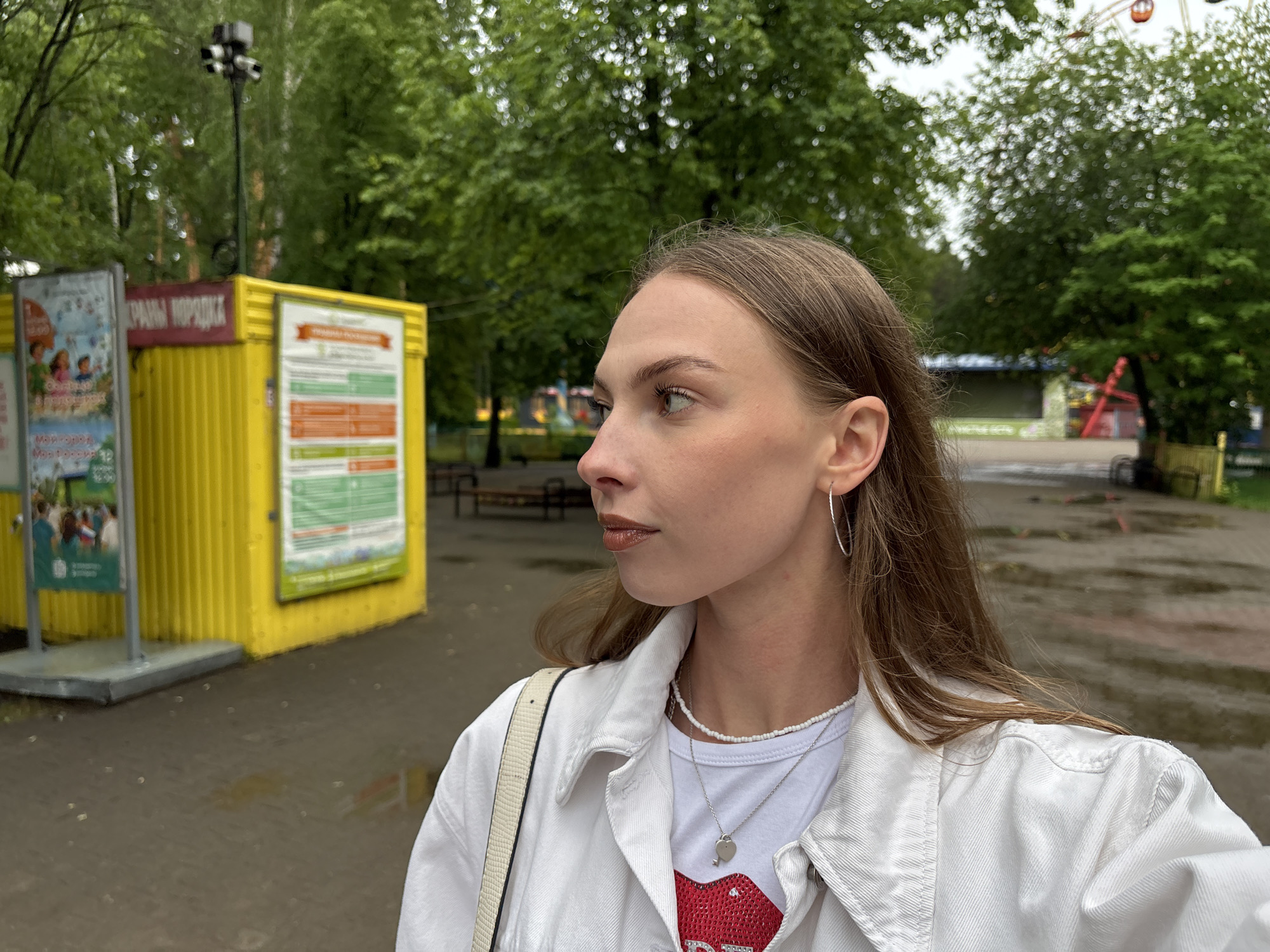 The image depicts a young adult female in a side profile, looking towards the left, within an outdoor park setting in Perm, Russia. She has long, light brown hair, visible brown eyes, and is wearing a white collared denim jacket over a white t-shirt. Her visible accessories include a short white beaded necklace, a longer silver chain with a heart-shaped pendant and a small key charm, and silver hoop earrings on her left ear. A light-colored bag strap is visible over her right shoulder.
The ground is a wet, dark paved surface with visible puddles, indicating recent rain. In the mid-ground, to the left of the woman, stands a bright yellow corrugated metal structure, appearing to be a kiosk or small building. Adjacent to it on the left is a grey stand displaying a poster with images of children and partially visible text, including "12:00." Mounted atop the yellow structure is a red sign with Russian text reading "ХРАНЫ ПОРЯДКА" (Guards of Order/Law Enforcement), and a tall pole equipped with multiple surveillance cameras.
The background consists of dense green deciduous trees. Visible above the tree line to the far right is a Ferris wheel with orange gondolas, and to its left, a red amusement park ride structure. The sky is overcast.