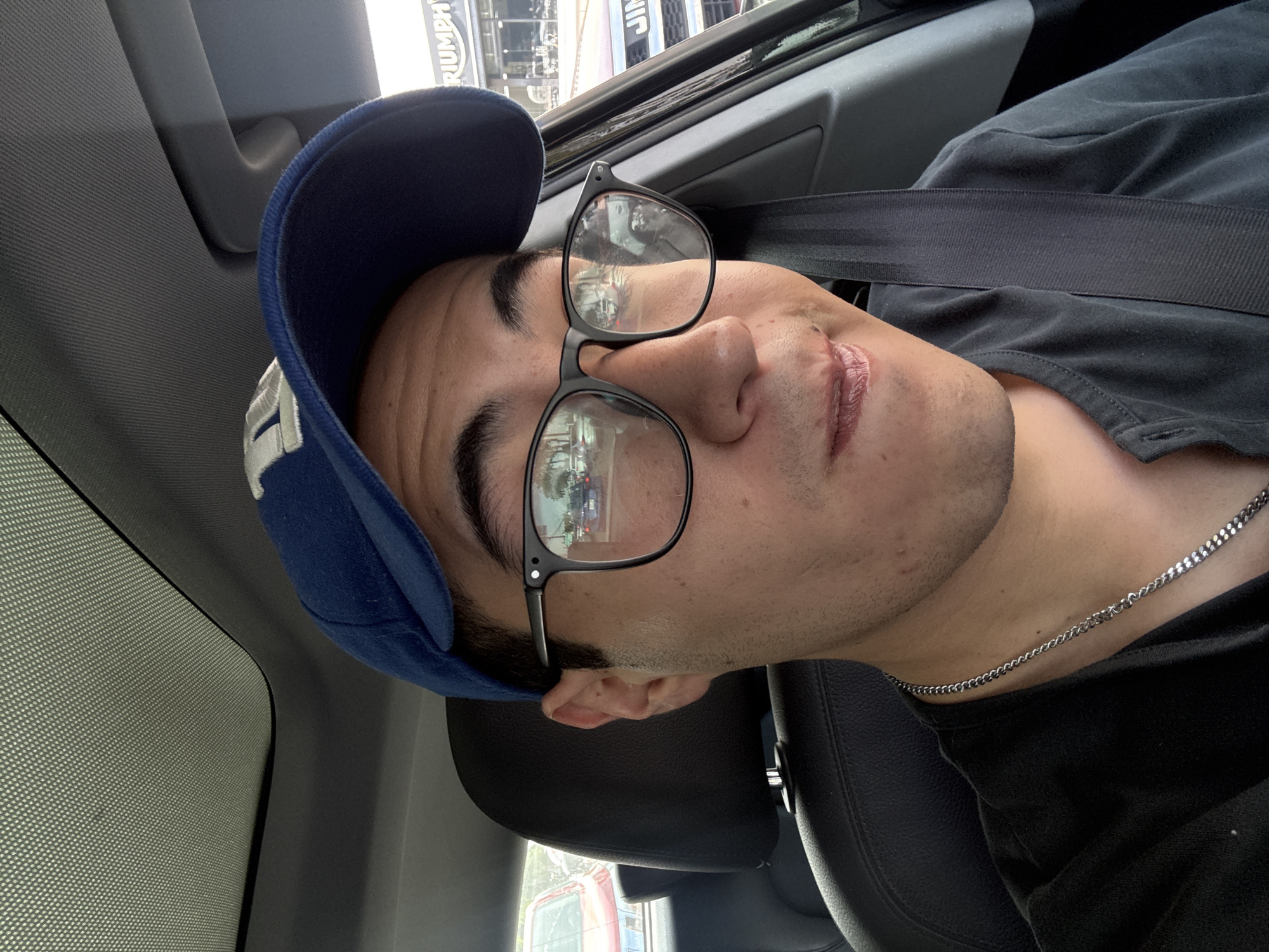 A close-up, angled portrait features a young adult male seated inside a car, likely captured as a selfie. He has light skin, dark eyebrows, and is wearing a dark blue baseball cap with a white logo on the side. He sports black-rimmed glasses, a black t-shirt, and a silver chain necklace. His lips appear somewhat dry or chapped, and he has light stubble. His expression is neutral as he looks directly towards the camera.
The interior of the car is visible, including a dark gray headliner, a grab handle, and a black seatbelt positioned across his chest. A black car seat or headrest is also partially visible behind his ear. The lighting suggests it is daytime, with diffused light entering the vehicle.
In the reflections of his glasses and through the car window, parts of the external environment are visible. A prominent white sign with the word "TRIUMPH" in dark letters is clearly discernible, indicating a commercial establishment, possibly a dealership or store. This provides a specific location cue within the broader context of Puebla, Mexico.
