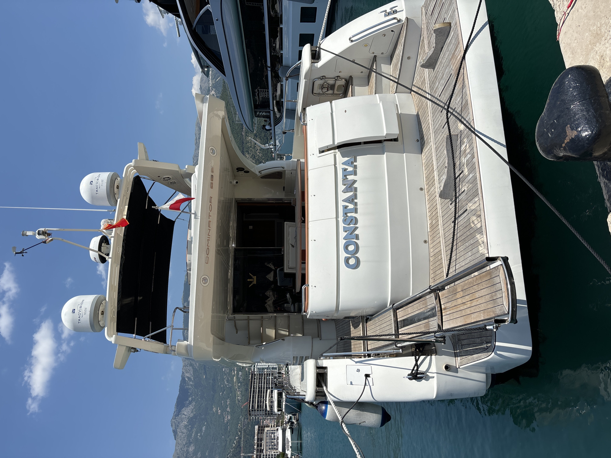 A large white yacht named "CONSTANTIA" is docked at a marina in Budva, Montenegro, under a clear blue sky with scattered clouds. The yacht features two large satellite domes on its upper deck, one labeled "TRACVISION" and "KVH". Two small flags, one red and one white, are attached to the mast. The stern of the yacht displays a wooden deck and a retractable swim ladder. In the background, other yachts are visible, as well as a city skyline with apartment buildings and cranes. Mountains form the backdrop to the scene.