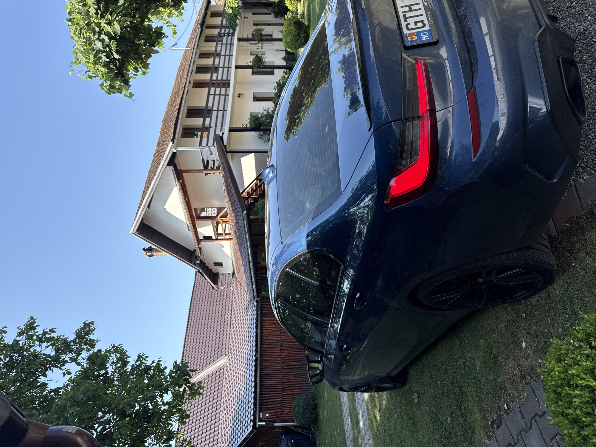 The image depicts a dark blue sedan parked on a grassy area in front of a two-story white building with a brown tiled roof. The building has balconies with wooden railings and several windows. A wooden fence is visible to the left of the building, and trees with green foliage are present in the foreground and background. The car's license plate, partially visible, reads "GTH" with a Moldovan flag emblem. The sky is clear and blue. The location is identified as Cârțișoara, Romania.