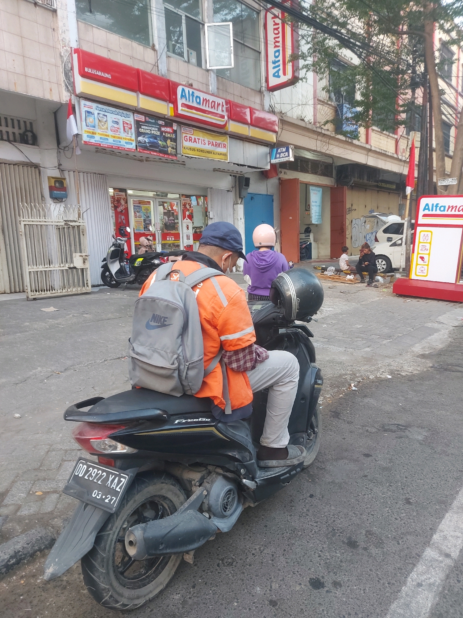 In Makassar, Indonesia, an outdoor scene unfolds in front of an Alfamart convenience store. A person wearing an orange jacket, a blue cap, and light-colored pants sits on a black scooter, facing away from the camera. The scooter has a license plate that reads "DD 2922 XAZ" and "03-27". The person has a gray Nike backpack on. To the left of the scooter, a metal gate is visible. In the background, the Alfamart store has various advertisements and an ATM BCA. Further in the background, another person wearing a purple hoodie and a pink helmet is also on a scooter. A white car and a few other individuals are present near the store entrance. The environment appears to be a street with cobblestone pavement.