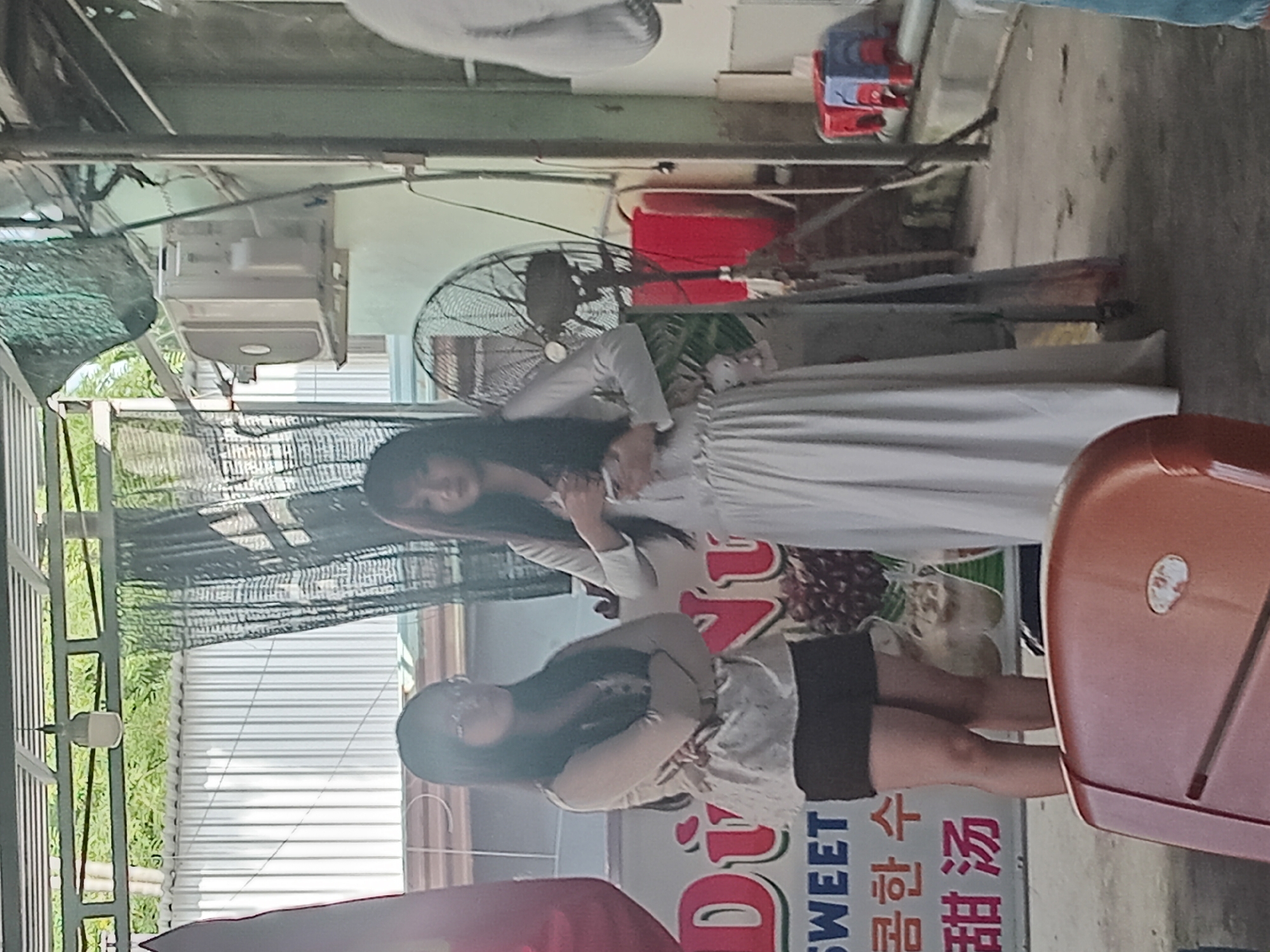 Two young women stand in an outdoor covered area. The woman on the left, wearing glasses, a light-colored blouse, and dark shorts, has her arms crossed. The woman on the right, in a long, flowing white dress, holds her hands near her chest. Behind them, a sign with text in English, Korean, and Chinese is visible, along with a large fan and an air conditioning unit. The ground is concrete, and there are various objects in the background, including red containers and a metal stand. The overall setting appears to be a street-level eatery or stall in Hoi An, Vietnam.