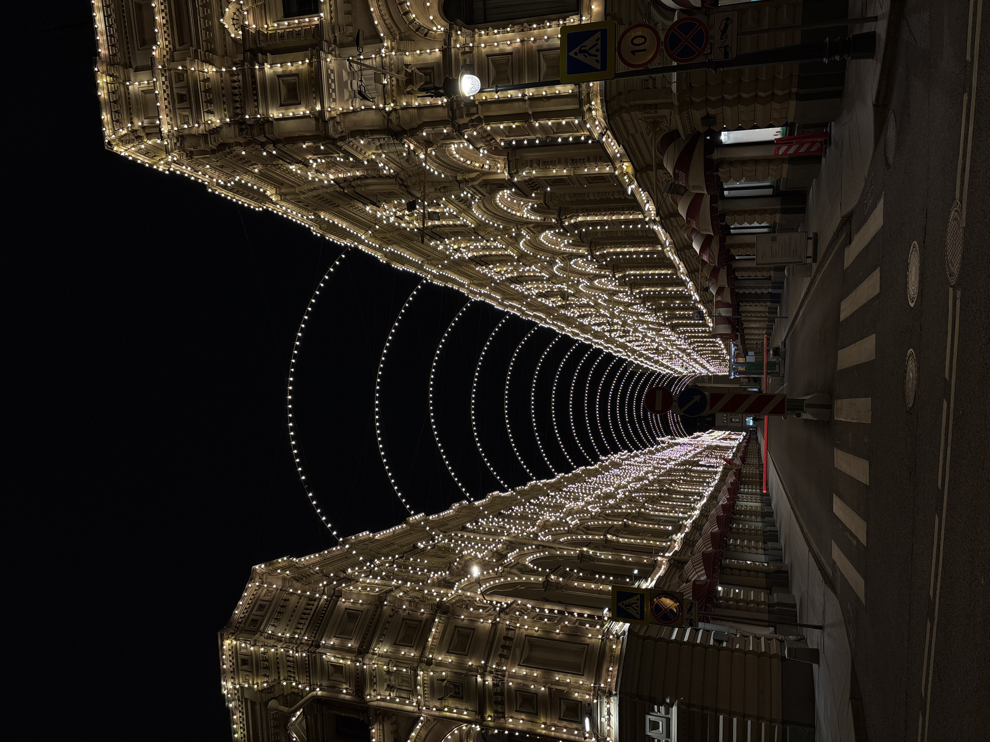 The image shows a street at night in an unknown location, decorated with extensive lighting. The buildings on either side are adorned with numerous small, warm-toned lights, outlining their architectural details. Strings of lights are hung across the street, forming illuminated arches. A pedestrian crossing is visible on the right side of the frame, marking the intersection of the street. Some traffic signs are visible on the side of the building. The background is dark, highlighting the contrast of the illuminated buildings.