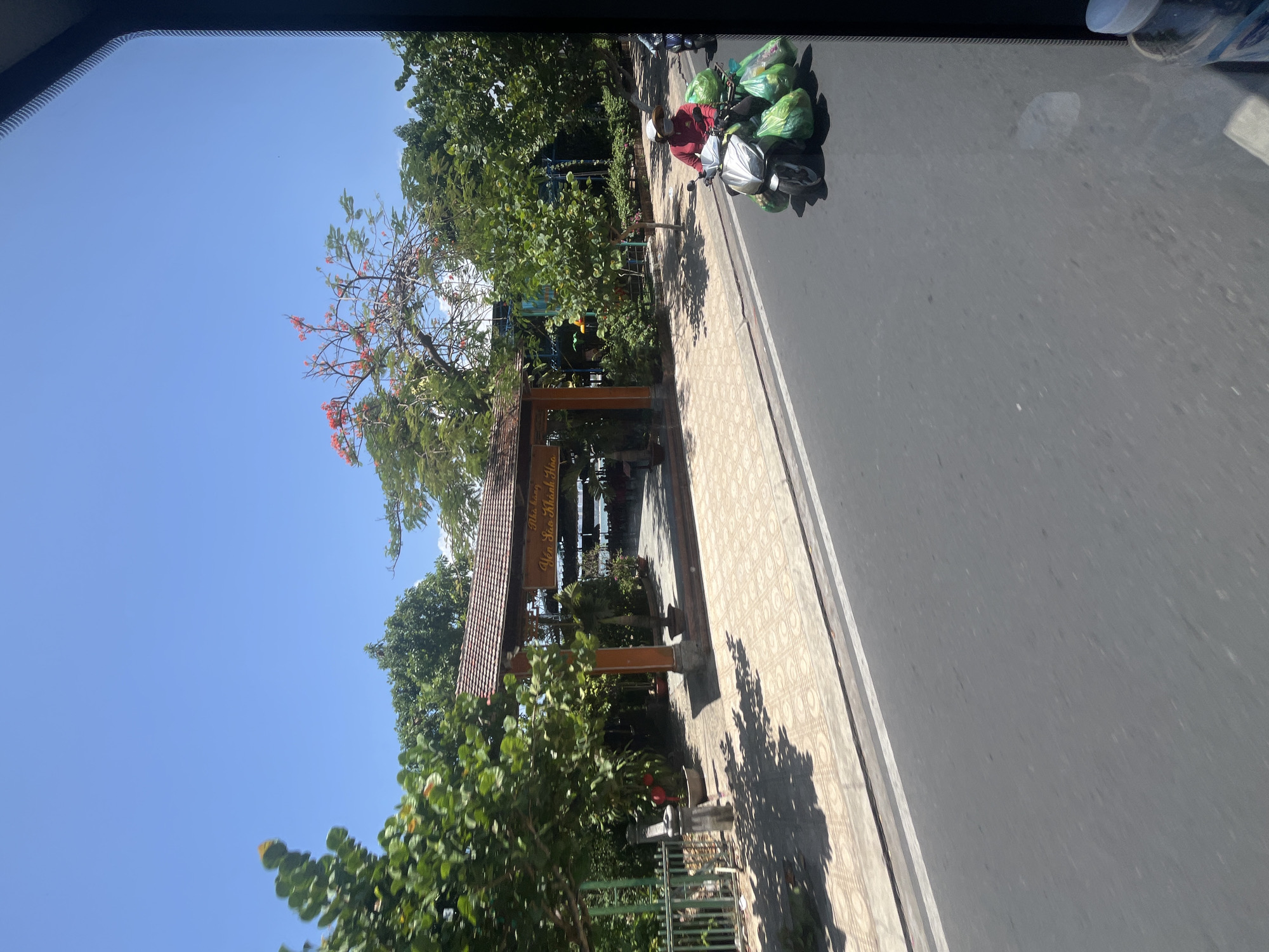 The image shows a street scene in Nha Trang, Vietnam, on a bright, sunny day. The foreground is dominated by a paved road and sidewalk. A person wearing a red long-sleeved shirt, a white hat, and a face mask rides a silver scooter loaded with numerous green plastic bags, suggesting they are transporting goods. The scooter is positioned on the road, to the right of the sidewalk.
Behind the sidewalk is an entrance to a building with a traditional wooden roof and decorative wooden posts. A sign with Vietnamese text, "Nhà hàng Yến Sào Khánh Hòa," is visible above the entrance, indicating it is a restaurant specializing in swiftlet nest products. Lush green trees with some red flowers are abundant, framing the entrance and the scene. The background shows more greenery and a glimpse of what appears to be a body of water or a distant view through the trees, likely part of the restaurant's setting. The sky is a clear, vibrant blue, indicative of a warm day. The perspective suggests the photo was taken from a moving vehicle, likely a car or bus, looking out towards the street.