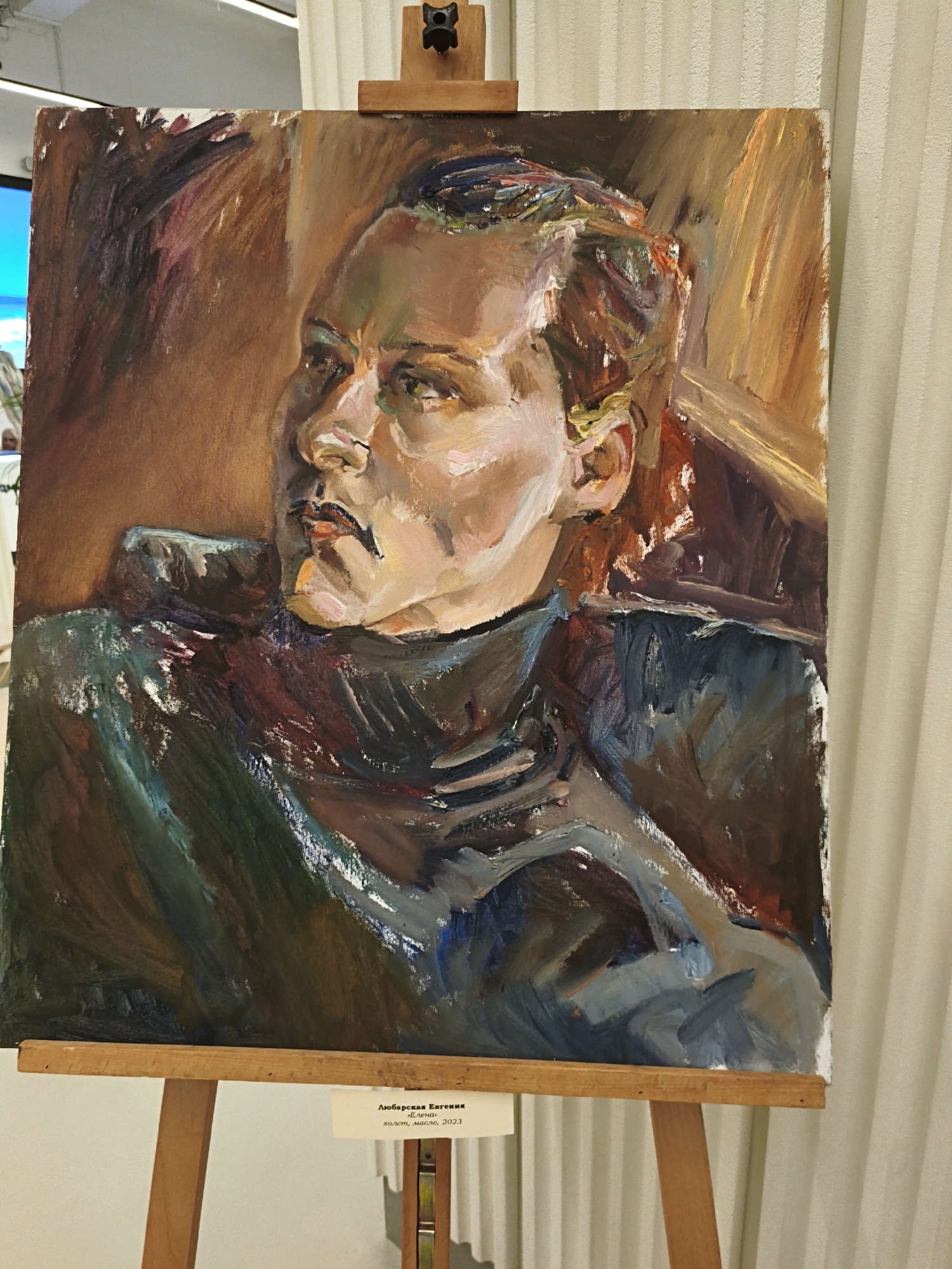 The image shows an oil painting on canvas, propped on a wooden easel. The painting depicts a portrait of a person with light skin and dark hair, looking slightly to the left. The person is wearing a dark-colored, high-necked garment. The background is a blend of brown and ochre tones, suggestive of an indoor space. The style of the painting is expressive, with visible brushstrokes and a focus on light and shadow. A small white label is attached to the easel beneath the painting; the text on the label is in Cyrillic. The background is blurred, showing a white wall with vertical texture. The location, Sevastopol, Ukraine, is not visually discernible in this image.
