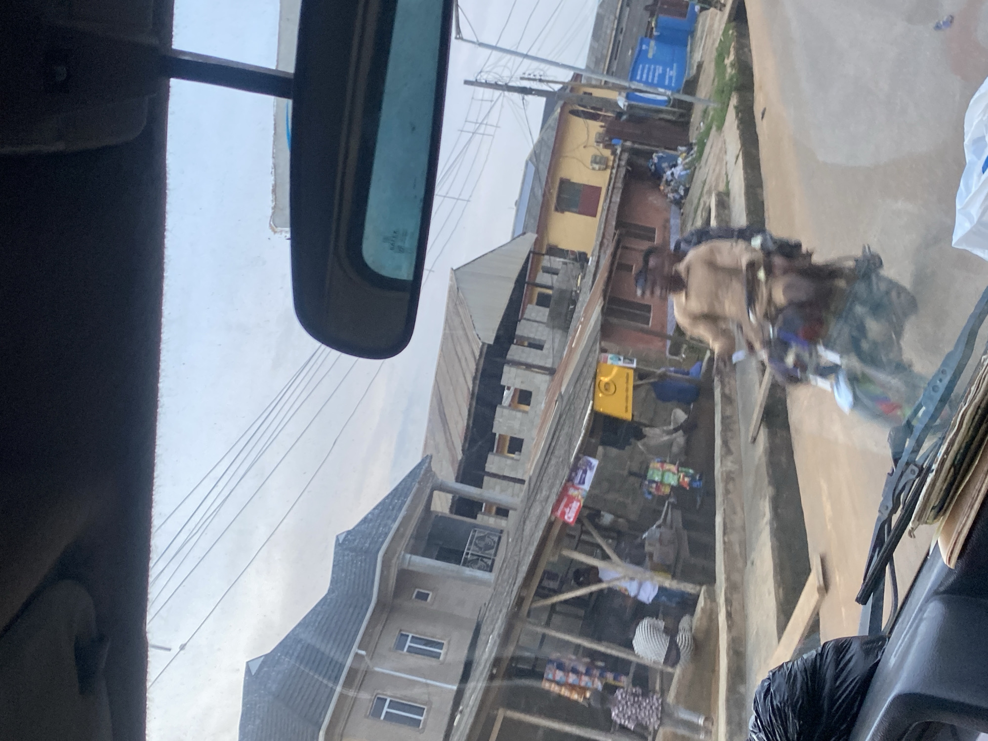 In Ikorodu, Nigeria, a motorcycle is seen traveling on a street lined with buildings and small shops. Two men are on the motorcycle, with the driver wearing a light brown shirt. In the background, several buildings of varying architectural styles are visible, some appearing to be residential and others commercial. Wooden structures, likely shops or stalls, are situated along the street, displaying various goods. A few other individuals are seen walking or standing near the shops. The street itself appears to be paved, with a drainage channel running alongside it. Power lines are strung overhead. The sky is overcast, suggesting it may be an early morning or late afternoon.