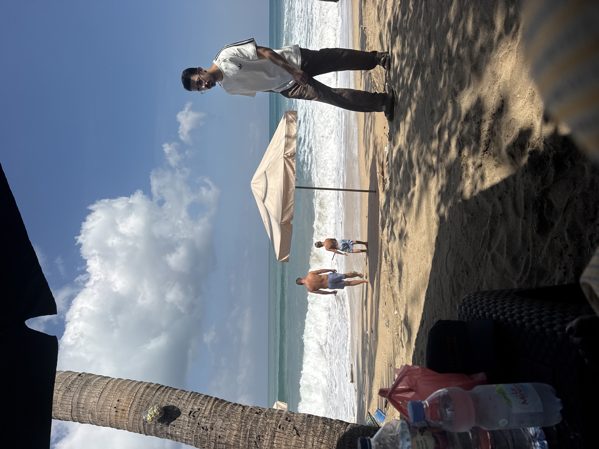 The image shows a beach scene in Kuta, Indonesia, on a sunny day with a clear blue sky and some clouds. In the foreground, a partially visible table with water bottles and a red bag are on the sand, casting a shadow. A person is seated in the lower right corner, mostly out of frame. Further back, a man in a white t-shirt and brown pants stands near a beach umbrella. Two other figures, a man and a boy, are walking towards the ocean, where large waves are breaking on the shore. A palm tree is visible on the left side of the image.