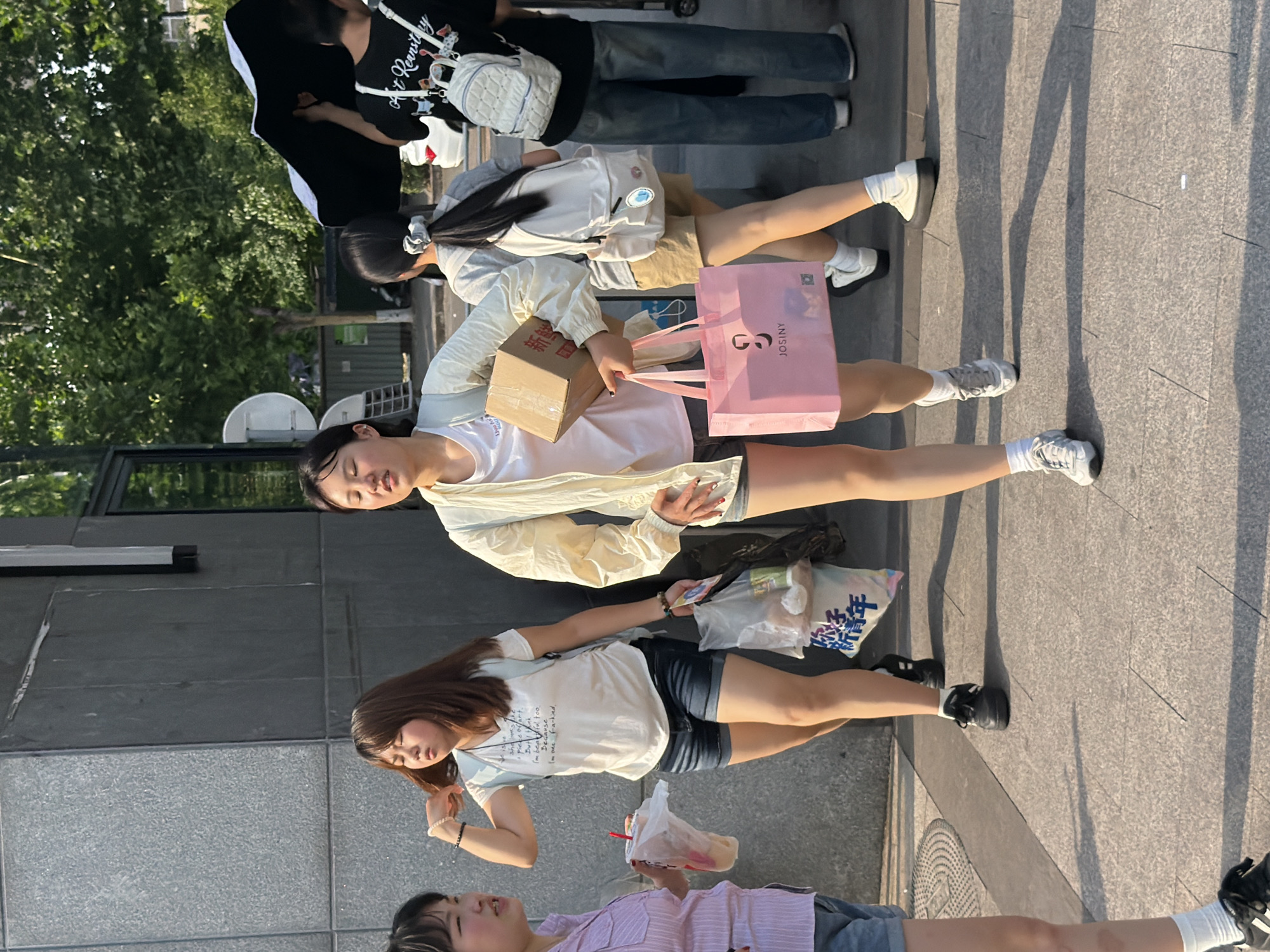 The image captures a street scene in Jiangjin, China, featuring several young women walking along a gray-tiled sidewalk on a sunny day. A group of four individuals are visible at the top of the frame, with the first person wearing a black shirt with white text and graphics, paired with light blue jeans. They have a white bag over their shoulder. To their right, a woman with a ponytail wears a light-colored jacket and khaki shorts.
Further down the frame, a woman in a white t-shirt and shorts carries a brown cardboard box and a pink shopping bag with the word "JOSINY" printed on it. To her right, another woman in a white t-shirt and dark shorts holds a clear plastic bag with Chinese writing. She also has a bracelet on her left wrist.
Below them, a woman in a light purple t-shirt and gray shorts holds a beverage in a plastic cup with a straw.
The sidewalk and building exteriors are visible in the background. Shadows of pedestrians and trees stretch across the sidewalk, creating depth in the image.