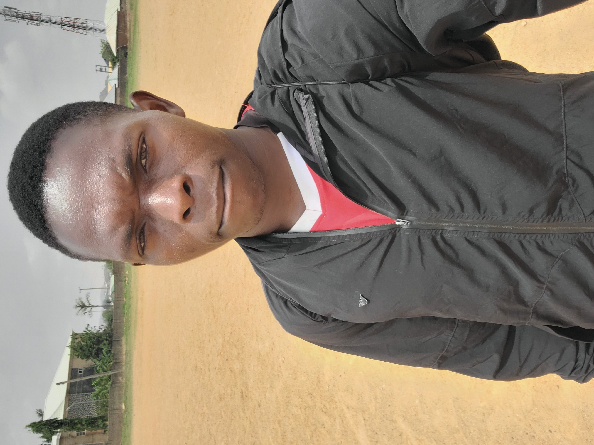 The photograph shows a man wearing a black jacket and a red and white shirt. He appears to be standing outdoors on a dirt or sandy field. In the background, there are buildings, trees, and a telecommunications tower. The sky appears overcast. The location is likely in or near Bukuru, Nigeria, based on the prompt.