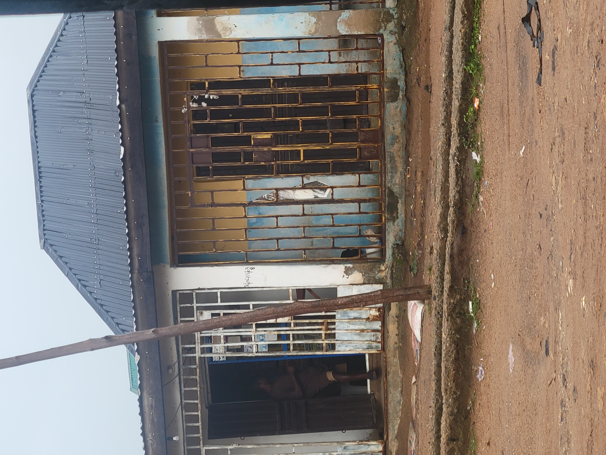 A medium shot captures a weathered building facade, likely a shop or residence, in Madalla, Nigeria. The building has a corrugated metal roof and a facade painted in alternating bands of yellow and faded blue. A thick metal grate covers the upper windows, showing signs of rust and damage. Below this, another grate covers a storefront or entrance, revealing a dark interior.
A person is visible inside the shop, appearing to be a man with dark skin wearing shorts. He is partially obscured by the grate and the dim interior lighting, making it difficult to discern his exact activity. A rough, wooden beam runs horizontally across the lower portion of the frame, obscuring part of the storefront.
The ground in front of the building is a rough, dirt path with some patches of sparse green vegetation growing along a raised edge. Debris, including small pieces of paper and plastic, is scattered on the path. The overall impression is one of a somewhat run-down but functional establishment in an outdoor, possibly dusty or muddy, environment. There is no clear indication of the time of day or specific weather conditions, though the lighting suggests it might be overcast or late in the day. Visible text on the building, above the lower grate, appears to be a handwritten label: "TRIMAG/SS/B".