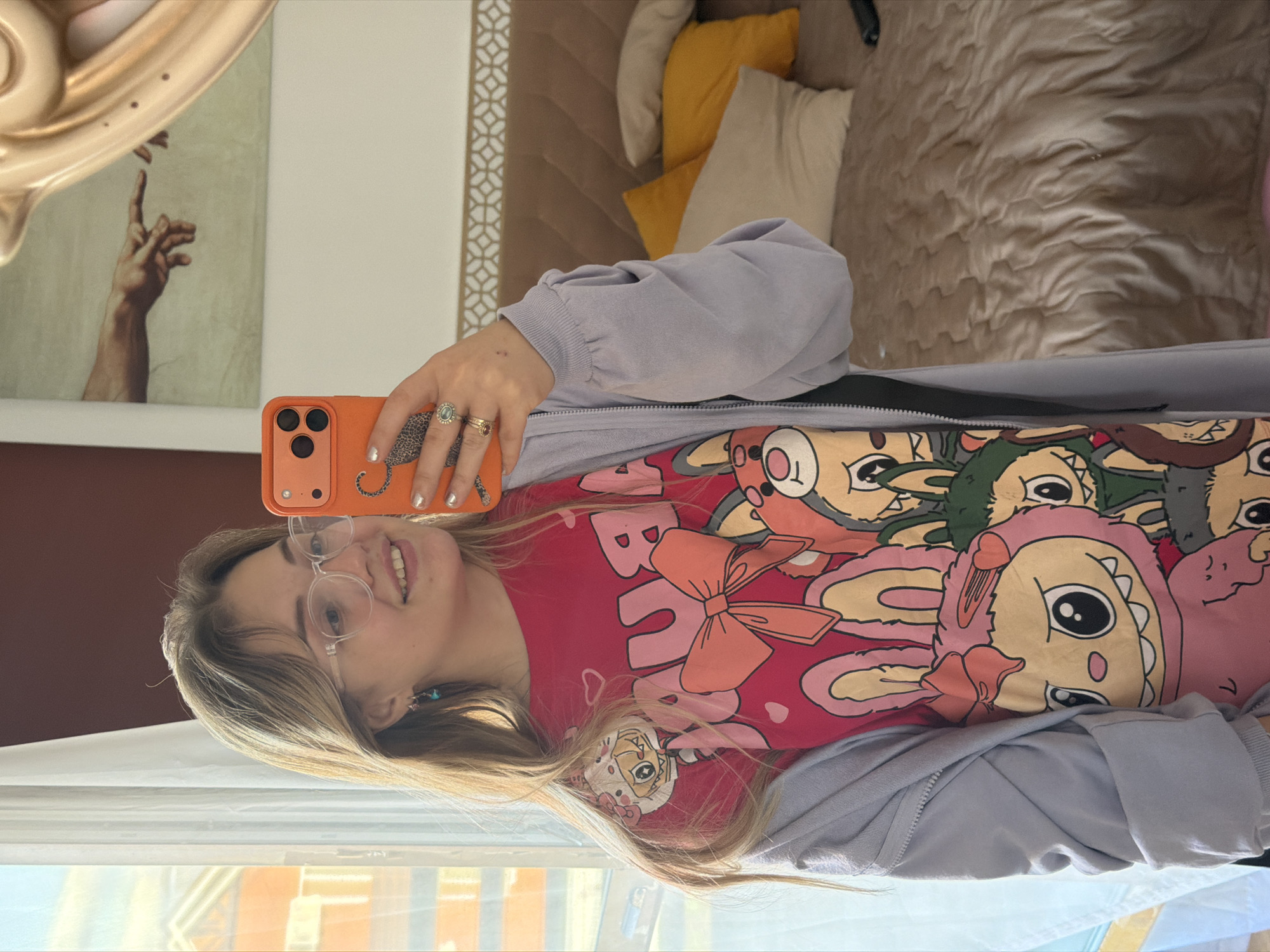 A young woman is taking a selfie indoors, smiling broadly at the camera. She has long, light-colored hair and wears clear-framed glasses. She is dressed in a vibrant red t-shirt featuring multiple cartoon characters, prominently displaying the word "BUNNY" and a large pink rabbit-like creature with a bow. Over her t-shirt, she wears a light purple or grey hooded jacket. On her fingers, she has several rings, including one with a large oval green or blue stone and other gold bands. Her nails are painted with silver polish, and a small blue earring is visible.
She holds an orange smartphone with three camera lenses in her right hand, part of its back cover showing a leopard print design. The background to her left features a framed artwork depicting a hand reaching out, reminiscent of Michelangelo's "The Creation of Adam." Above this, a golden, organic-shaped sculpture is partially visible.
To her right, a bed with a quilted brown headboard is visible, adorned with yellow, cream, and brown pillows. A decorative panel with a geometric pattern runs along the wall. Natural light streams in from a window in the foreground, reflecting parts of the outdoors. The image is presented rotated 90 degrees clockwise from its natural upright orientation. The scene suggests a relaxed daytime moment in what appears to be a bedroom or living area.
Location: Большая Шильна, Russia.