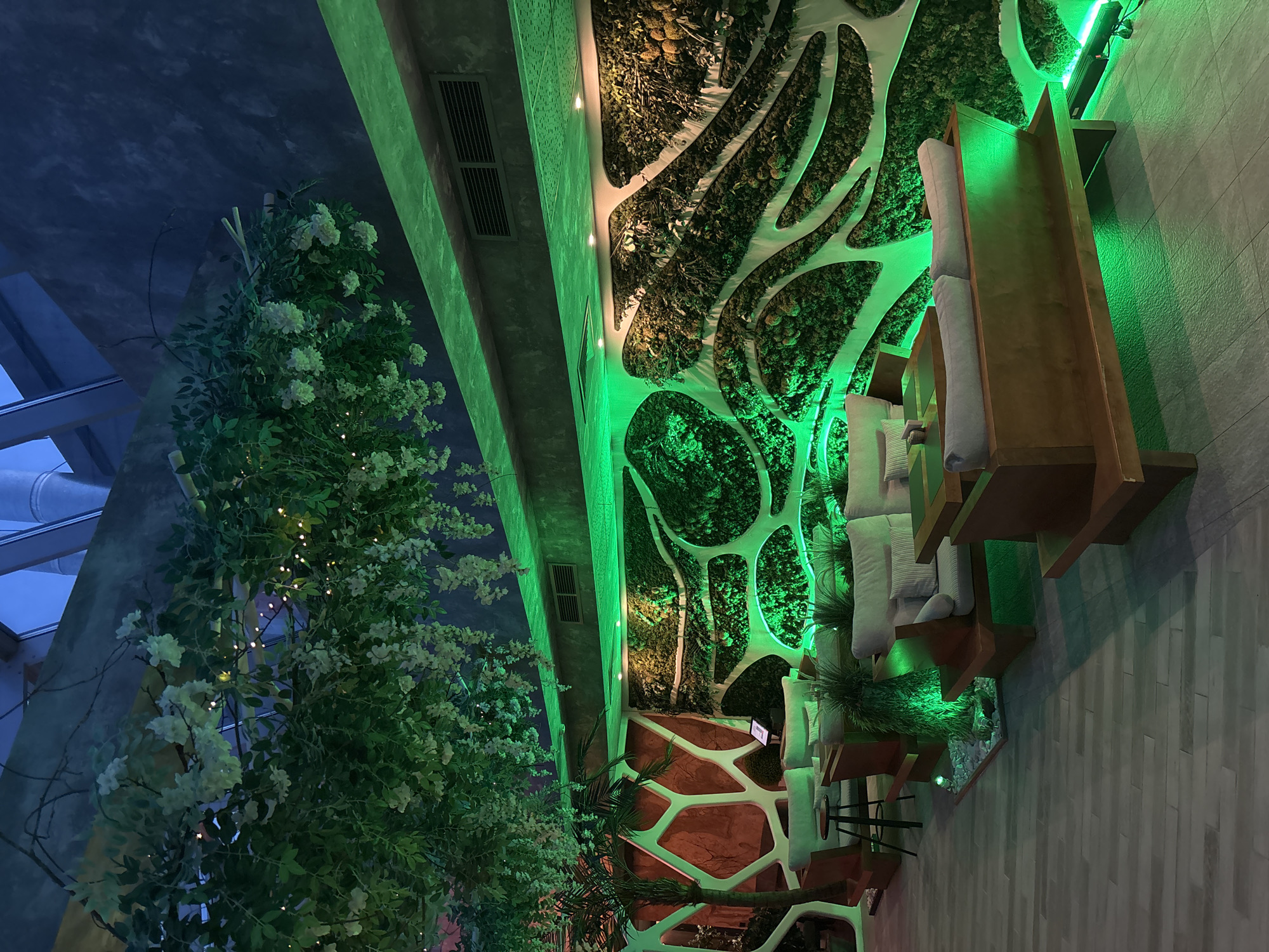 The interior of a restaurant in Minsk, Belarus is shown. The space is illuminated with green neon lights. A wall features a vertical garden interspersed with white geometric designs. A large flowering plant stands on the left, decorated with small lights. Seating consists of wooden-framed furniture with white cushions, including a high-backed bench. Tile flooring with a wood-like pattern runs along the right side of the image.