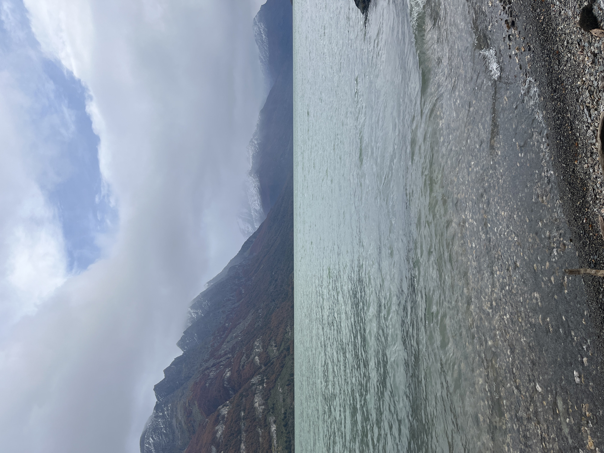 The image depicts a landscape near Lapataia, Argentina. On the left, there's a mountain range shrouded in clouds and mist, with some snow visible on the higher peaks. The mountain slopes are covered in vegetation, transitioning from green to reddish-brown. On the right, there's a body of water, possibly a lake or fjord, with a rocky shoreline covered in pebbles and stones. A small stick is visible on the right edge of the shoreline. The sky is overcast with a patch of blue peeking through the clouds on the left. No people, animals, or distinct landmarks are discernible in the image.