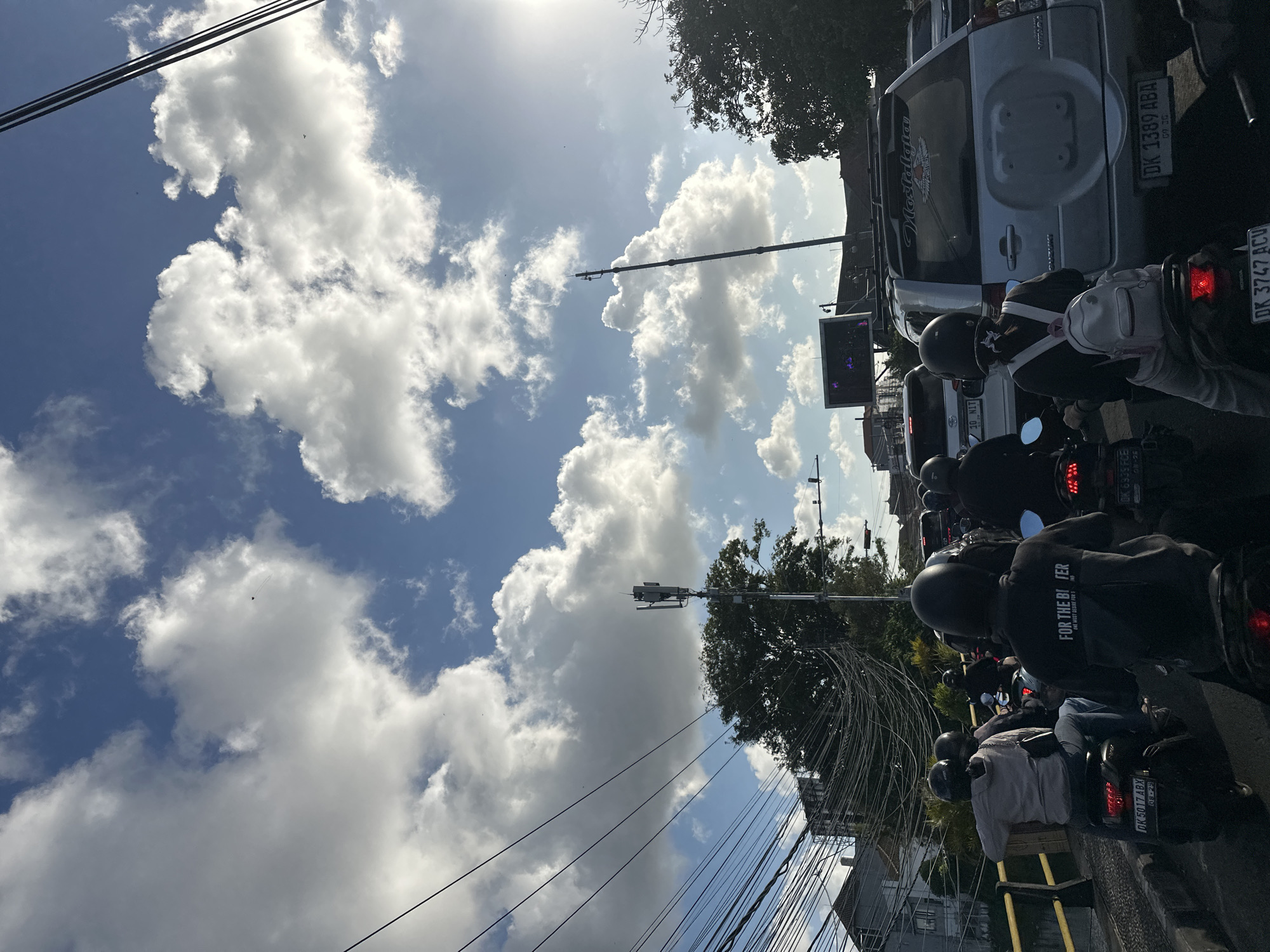 The image depicts a traffic scene in Denpasar, Indonesia, during the day. Numerous motorcycles and a few cars are stopped on a road, suggesting a traffic jam. People on the motorcycles are wearing helmets, with some passengers carrying backpacks. The environment is urban, with buildings and telephone poles visible. The sky is partly cloudy, with bright sunlight filtering through. There is a noticeable amount of electrical wiring overhead, a common sight in many Indonesian cities. The overall impression is one of a busy, bustling city street.