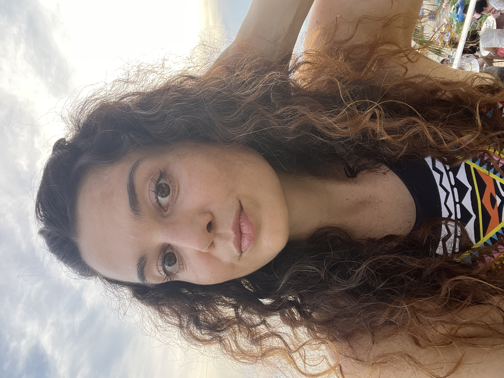 A woman with long, curly brown hair is posing for a selfie against a cloudy sky, likely at a beachside cafe in Mũi Né, Vietnam. The woman is looking directly at the camera, her right arm raised and hand touching her hair. She is wearing a black and patterned top. In the background, there are tables and chairs, suggesting an outdoor seating area, and hints of beach structures and foliage. The lighting indicates it is either sunrise or sunset.
