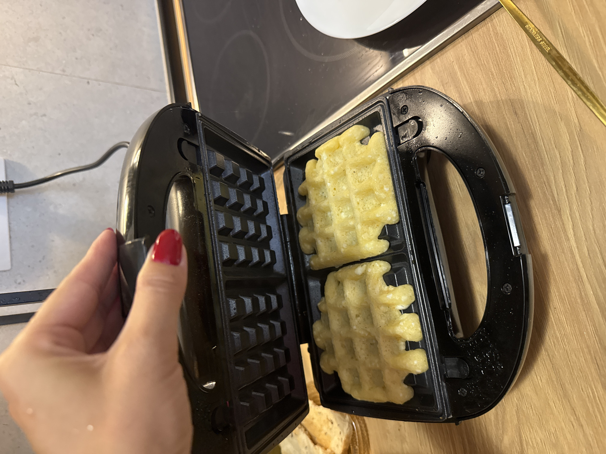 A hand with red fingernails is opening a black waffle maker. Inside the waffle maker are two golden-brown waffles. The waffle maker is on a wooden countertop, with a black stovetop and a white plate visible in the background.