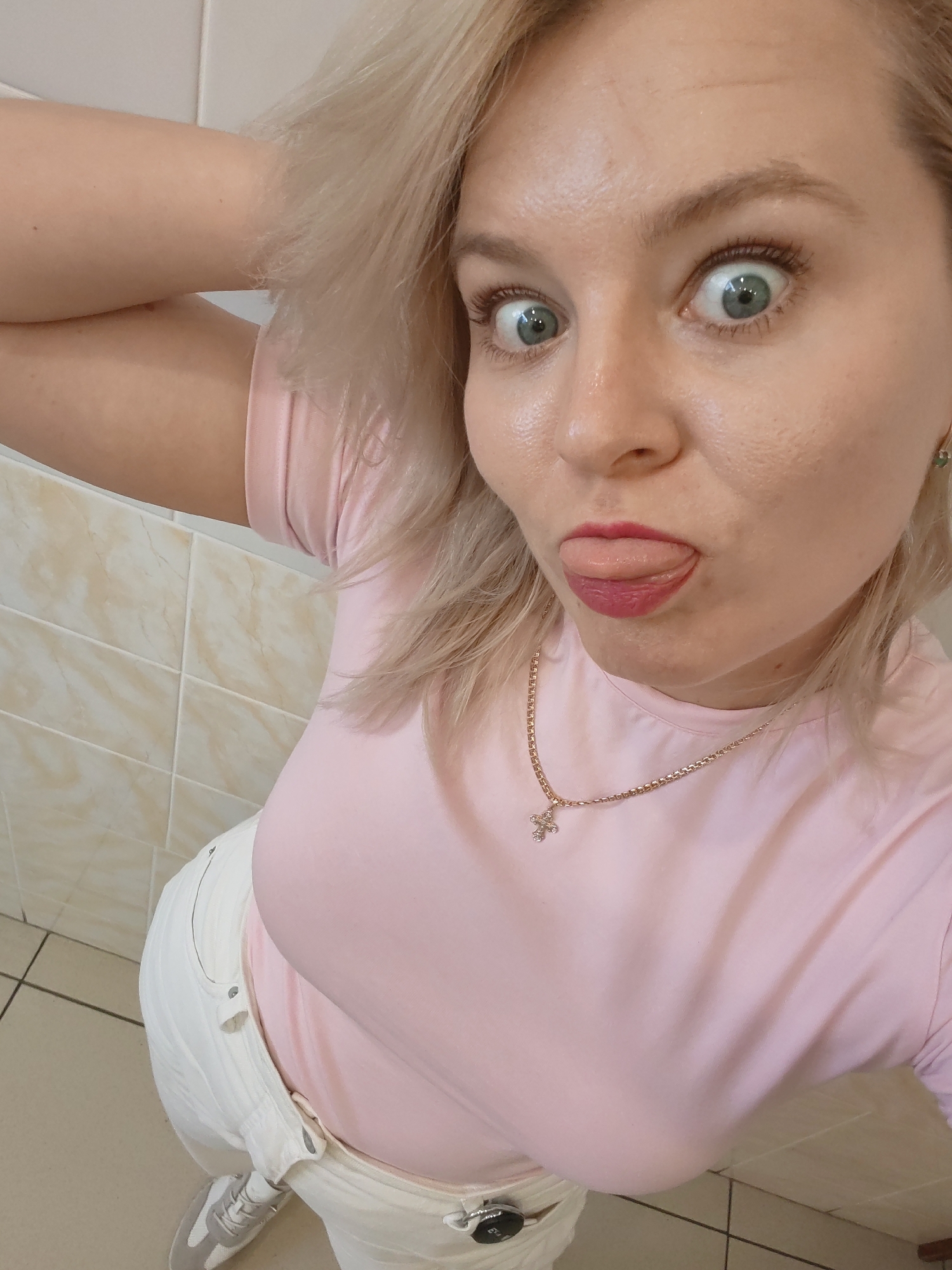A young Caucasian woman with blonde hair and blue eyes is taking a selfie. She is wearing a pink t-shirt, white pants, and grey sneakers. Her right arm is bent at the elbow, with her hand on her head. She is sticking her tongue out, and her eyes are wide, giving a playful or surprised expression. A gold necklace with a cross pendant is visible around her neck. The background appears to be a bathroom or tiled area, with beige tiles on the wall. The lighting suggests it is daytime.