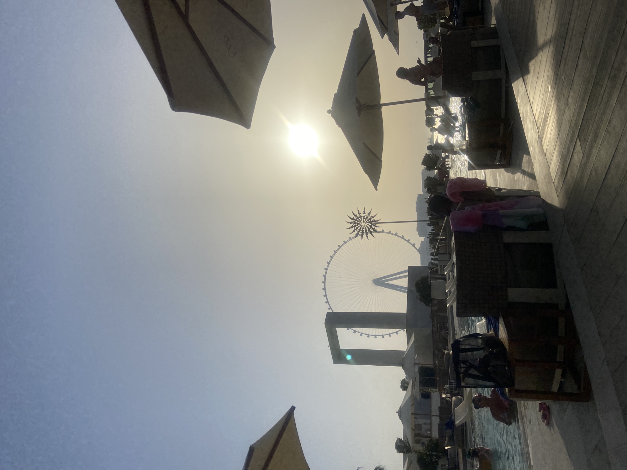 This image shows a view in Dubai, United Arab Emirates, near the Ain Dubai Ferris wheel. The scene includes beach umbrellas, a Ferris wheel, the beach, and beachgoers. People are swimming in the water. There is a wooden boardwalk or deck on the right side of the frame. The sun is high in the sky.