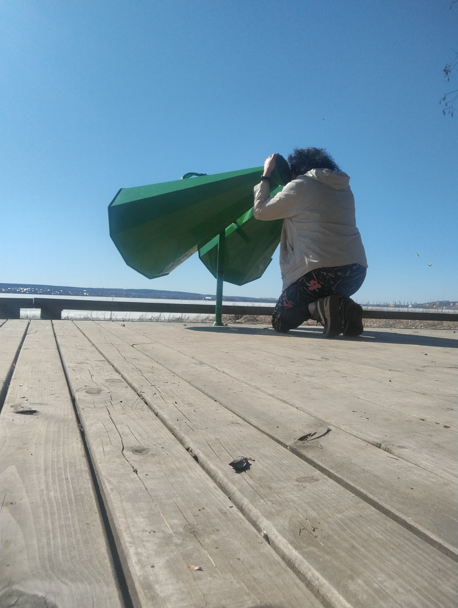 A person is kneeling and looking through a green observation telescope on a wooden platform. The person has dark, curly hair and is wearing a light-colored jacket and floral-patterned leggings. Beyond the platform, a body of water is visible with a distant shoreline. The sky is clear and blue. The location is in Votkinsk, Russia.
