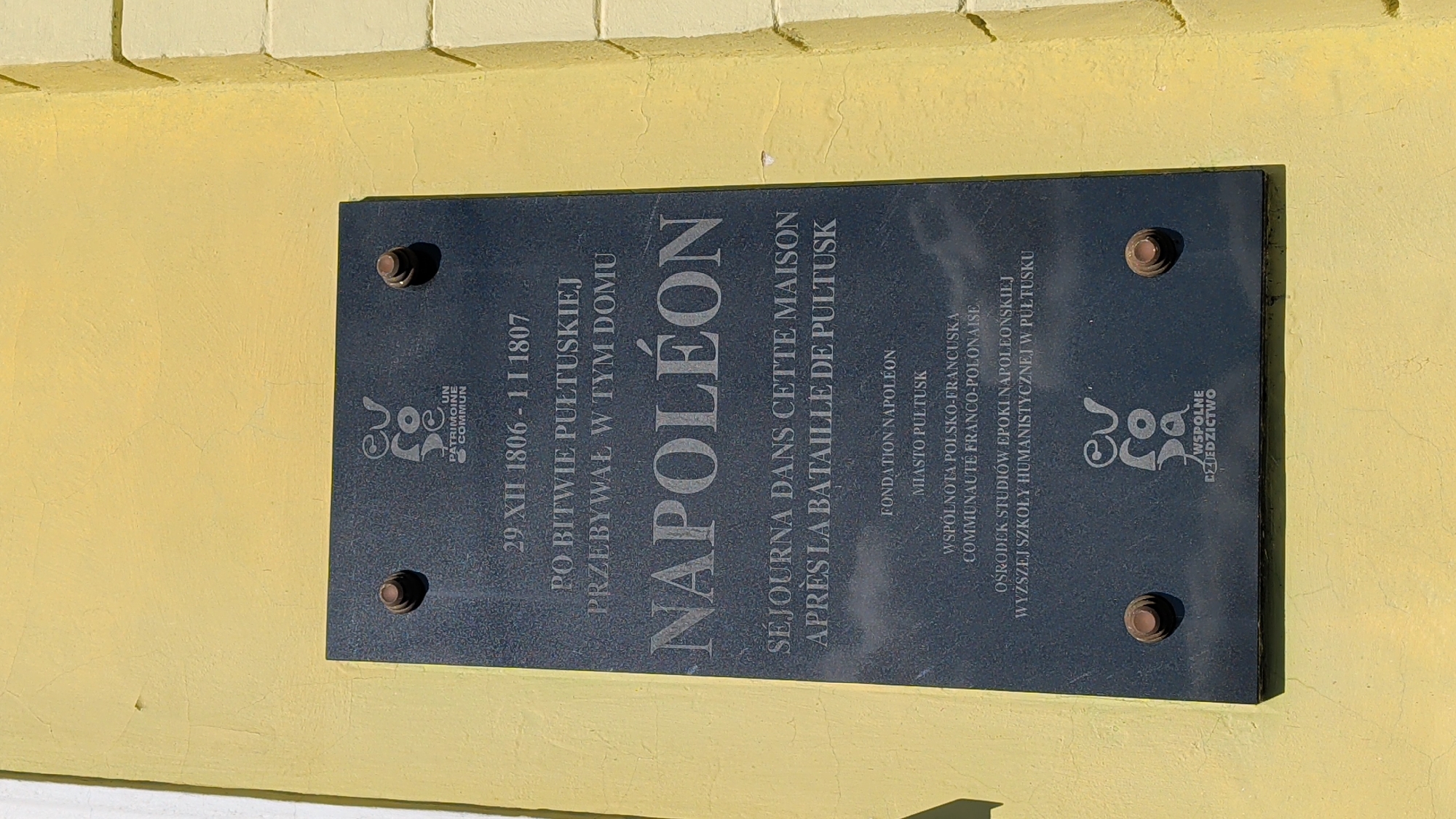 A dark granite plaque is mounted on a yellow building exterior. The plaque is engraved with text in both Polish and French. At the top center of the plaque is a circular logo with the letters "EU" intertwined with what appears to be a stylized "Ro" and "de" symbol, below which are the words "UN PATRIMOINE COMMUN". Below the logo, the plaque reads: "29 XII 1806 - 1 I 1807 PO BITWIE PUŁTUSKIEJ PRZEBYWAŁ W TYM DOMU NAPOLEÓN". This translates to "29 XII 1806 - 1 I 1807 AFTER THE BATTLE OF PUŁTUSK STAYED IN THIS HOUSE NAPOLEON". Further down, there is French text: "SÉJOURNA DANS CETTE MAISON APRÈS LA BATAILLE DE PULTUSK". Below this, smaller text indicates sponsoring organizations: "FONDATION NAPOLÉON", "MIASTO PUŁTUSK", "WSPÓLNOTA POLSKO-FRANCUSKA", "COMMUNAUTÉ FRANCO-POLONAISE", and "OŚRODEK STUDIÓW EPOKI NAPOLEOŃSKIEJ WYŻSZEJ SZKOŁY HUMANISTYCZNEJ W PUŁTUSKU". At the bottom of the plaque, the same circular logo appears again, with the word "WSPÓLNE DZIEDZICTWO" below it, translating to "COMMON HERITAGE". The plaque is secured to the wall by four visible screws with decorative heads. The building wall is painted a bright yellow with visible cracks in the plaster. The image is taken from a slightly upward angle, focusing on the plaque.