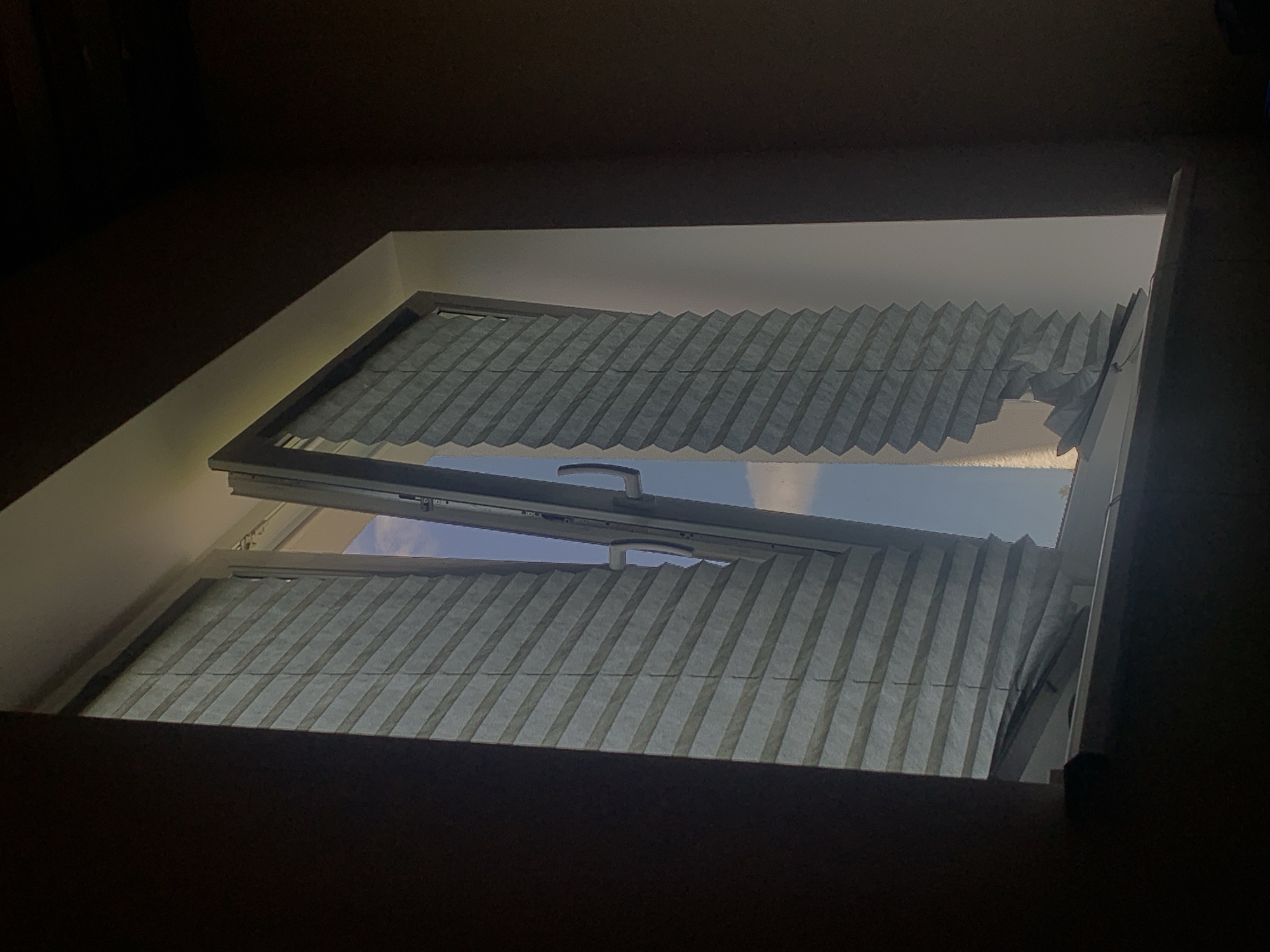 The image depicts a partially open window with light gray pleated blinds. The window frame is white, and the blinds are partially drawn, revealing a cloudy blue sky outside. The right blind is bent and damaged at the bottom, appearing torn or detached. The interior of the room is dimly lit, with dark walls on the right and left. The window sill is visible at the bottom of the frame. The scene is a domestic setting, possibly a residential apartment, and provides no specific information about the city of Minsk, Belarus.