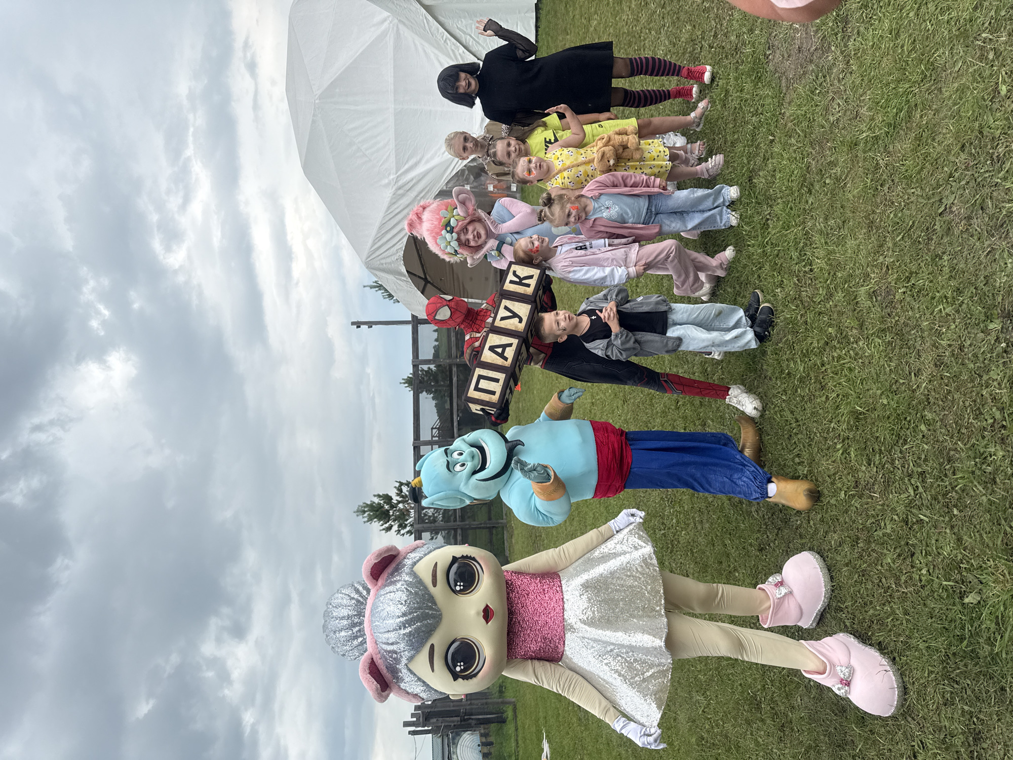 In a grassy outdoor area, likely on the outskirts of Kuznetskoye, Russia, a group of people are posing for a photograph. In the foreground, a character in a large, pink and silver costume resembling a cartoon cat stands to the left. To its right, a blue genie-like character from Aladdin is positioned, holding a wooden sign with Cyrillic letters that read "ПАУК". A person dressed as Spider-Man stands behind the genie and the sign. To the right of these characters, a line of children and two young women are arranged. One young woman has bright pink, troll-like hair and is positioned behind the children. Another woman, dressed in black with striped stockings, stands on the far right. A large, white, tent-like structure is visible in the background, suggesting an event or festival setting. The sky is overcast with clouds.