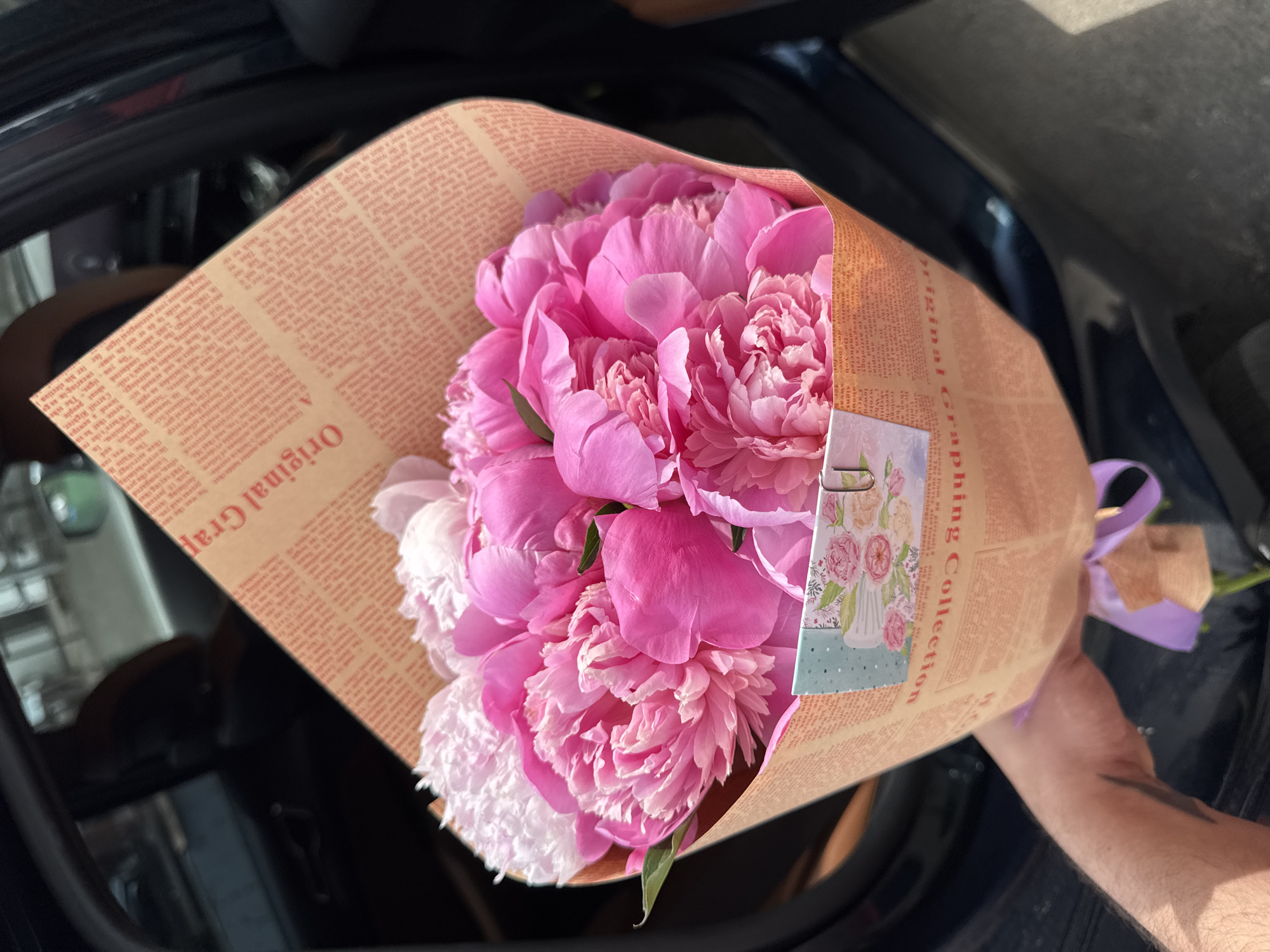 A person's arm is visible holding a bouquet of pink and white peonies wrapped in paper. The paper has a vintage newspaper print design with the words "Original Graphic Collection" visible. A small card with floral imagery is attached to the bouquet with a paperclip. The bouquet is positioned in front of a car interior, with dark upholstery and a dark exterior visible. The scene appears to be outdoors in daylight, with shadows indicating sunlight. There are no discernible landmarks or context to suggest the city of Almaty, Kazakhstan.