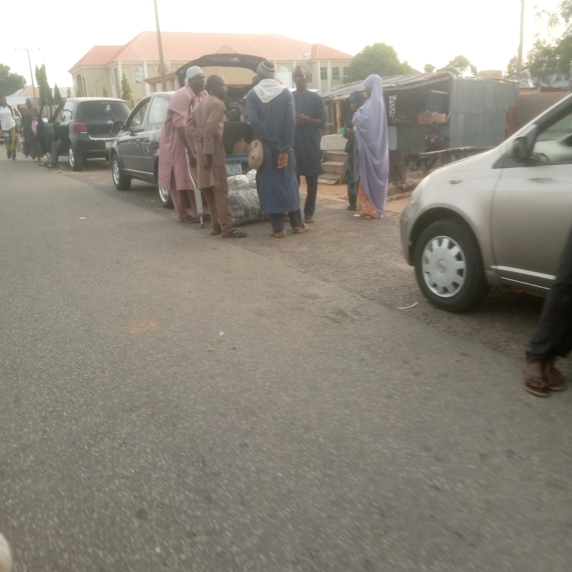 In an outdoor urban scene, several individuals are gathered near a row of parked cars and a corrugated metal structure that appears to be a small shop. The foreground is dominated by an asphalt road surface. In the mid-ground, a light-colored car is parked, its front wheel visible. To the left, a black car is also parked. A group of people, mostly men dressed in traditional clothing, are standing near the open trunk of one of the cars. Some are interacting with each other, while others appear to be observing. One person is wearing a blue garment and a headscarf, another a pink robe, and a third a brown robe. A woman in a light purple hijab stands further to the right, near the shop. The shop itself has a corrugated metal exterior and a wooden counter or display area. In the background, a multi-story building with a light-colored facade and an orange roof is visible, along with some trees. The sky is overcast. The scene suggests a street-side interaction or transaction, possibly involving goods being loaded or unloaded from vehicles, with the shop serving as a backdrop or point of commerce.
